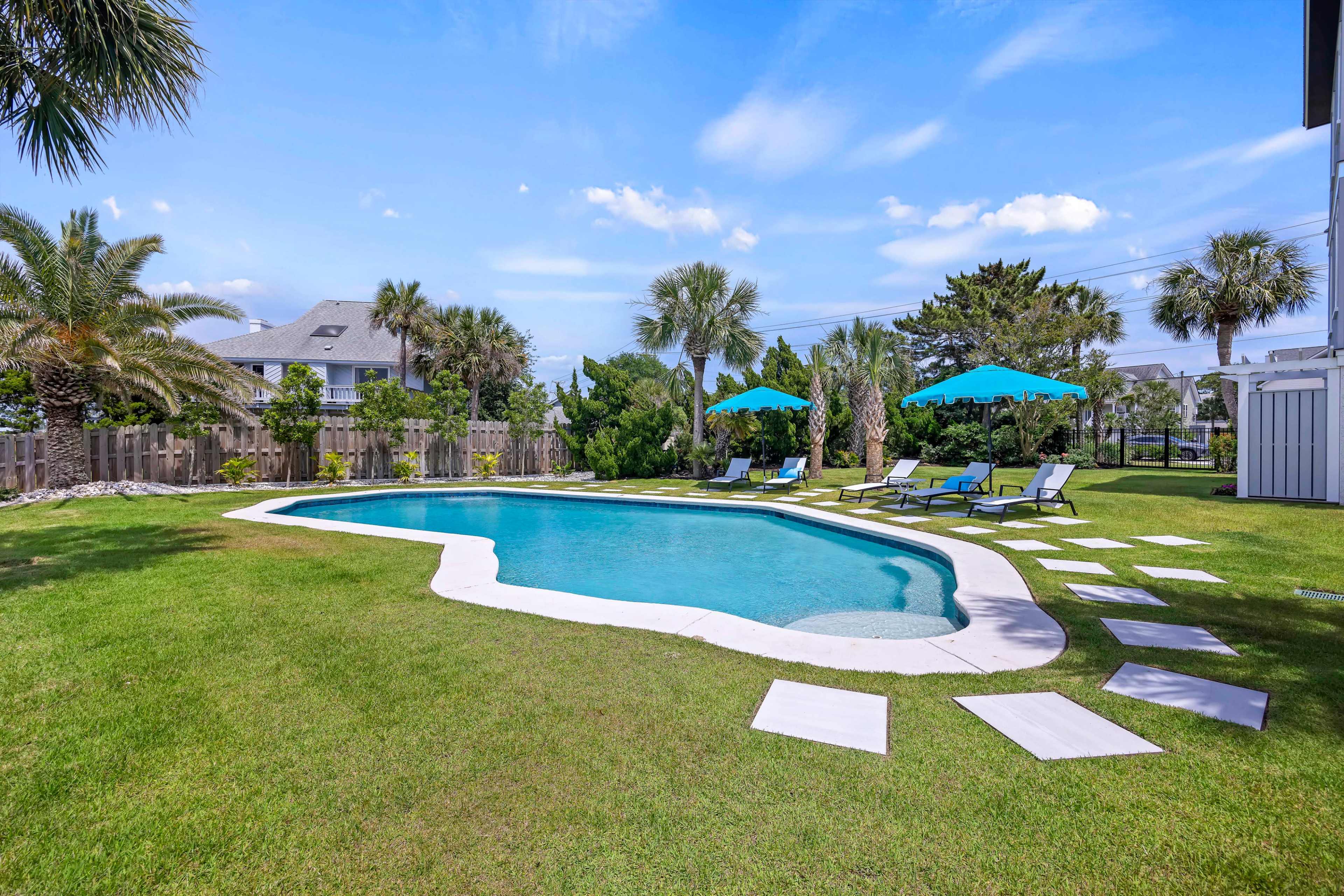 The image shows a landscaped backyard with a swimming pool surrounded by lounge chairs and umbrellas, along with neatly arranged stepping stones on the grass.