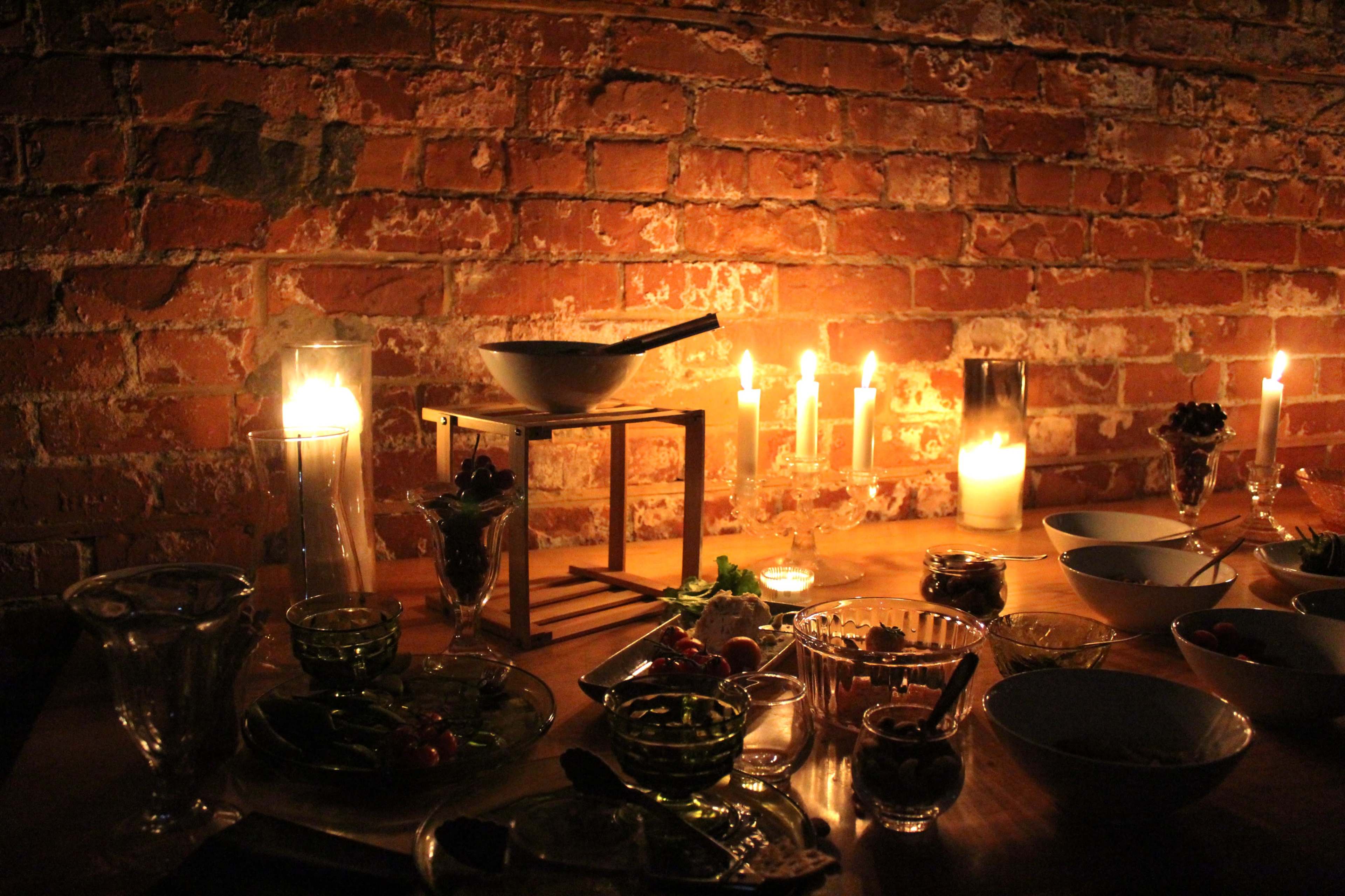 A wooden table is set with various dishes and glasses, illuminated by multiple candles and a warm light against a brick wall backdrop.