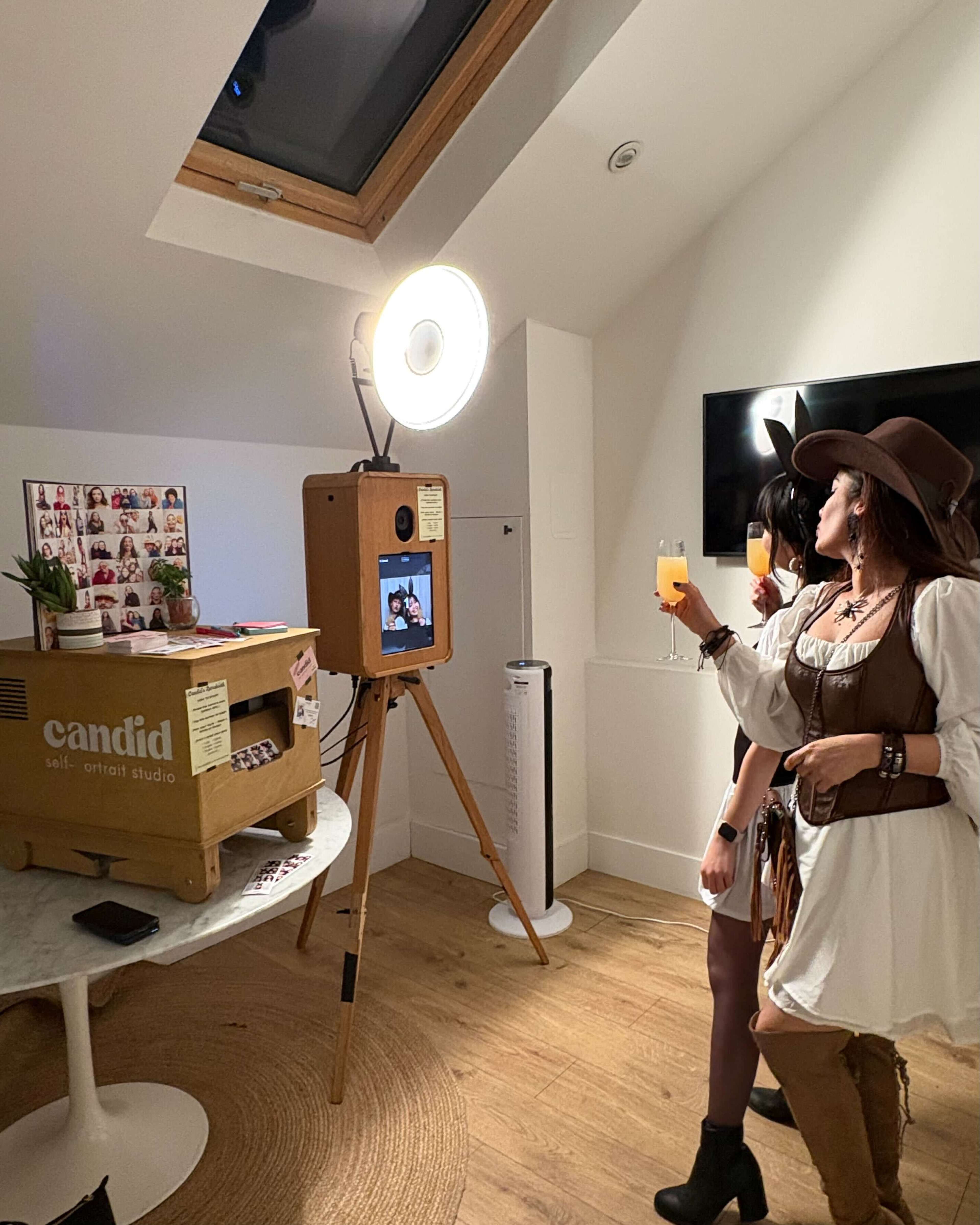 Two women dressed in Western-themed costumes hold drinks in front of a photo booth set up in a bright, modern room.