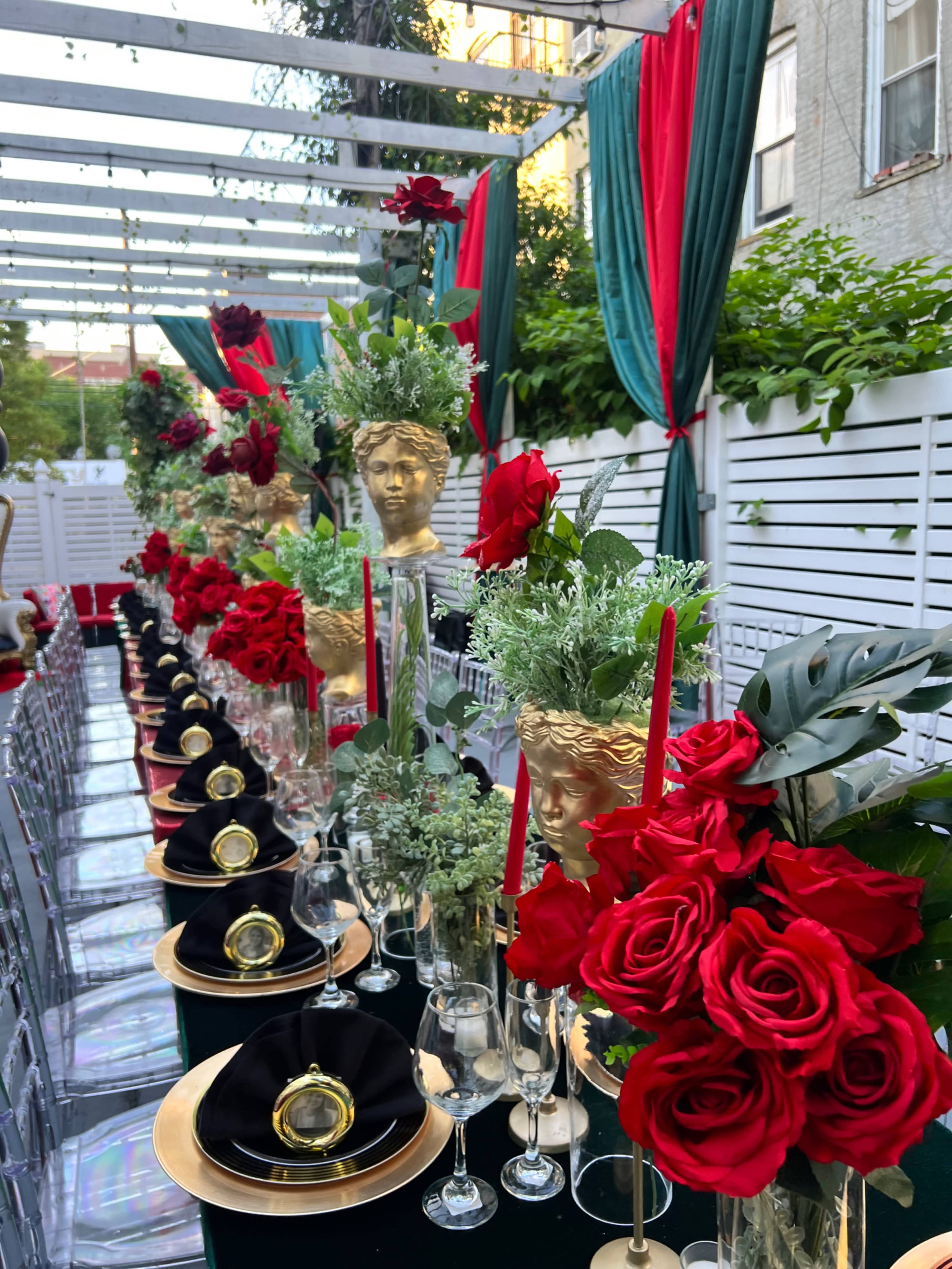 A long dining table is set outdoors, decorated with red roses, gold tableware, and ornate busts, under draped fabric in green and red.