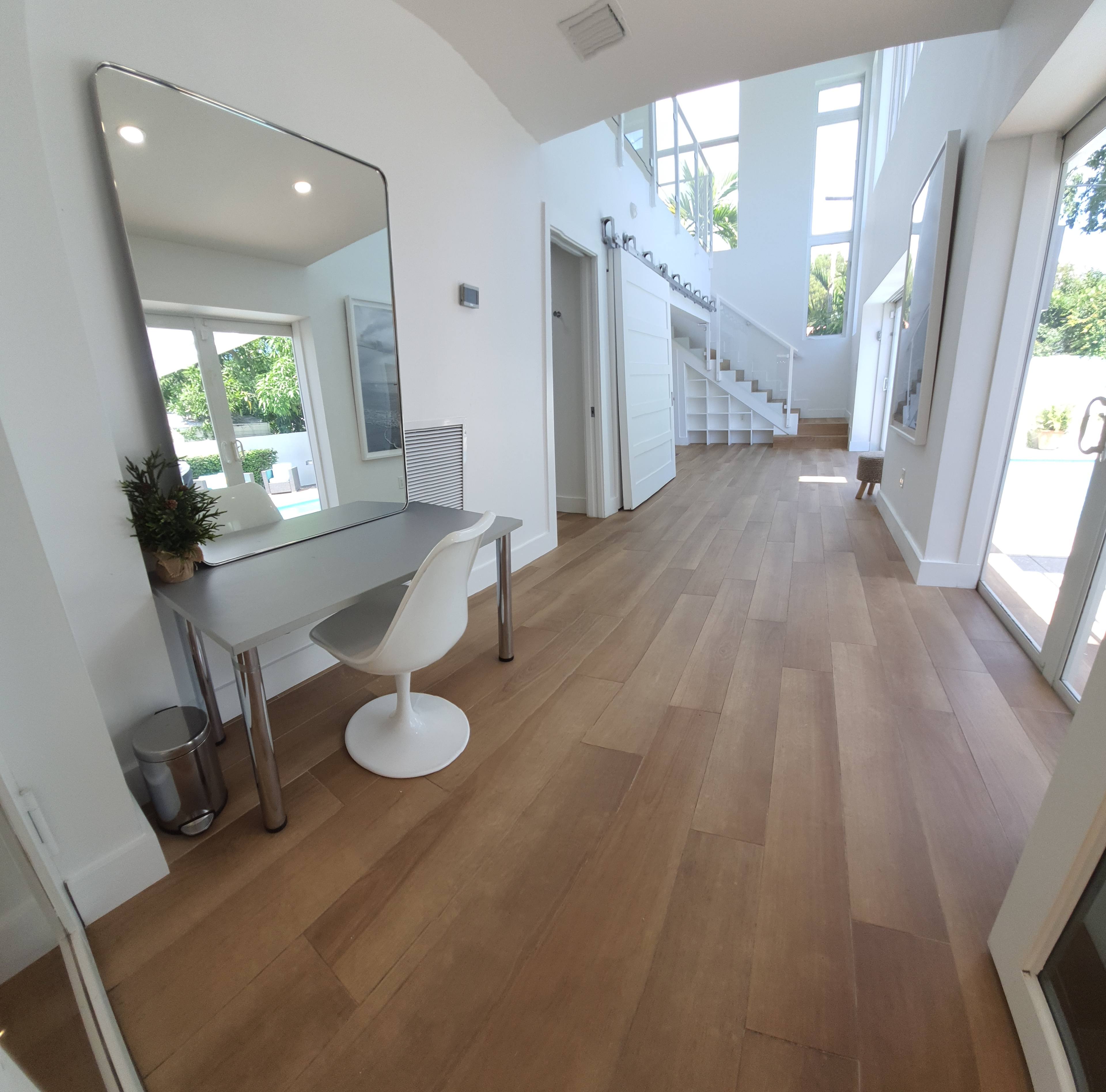 A spacious indoor hallway featuring a staircase on the right, a large mirror and a desk with a chair on the left, and large windows allowing natural light.