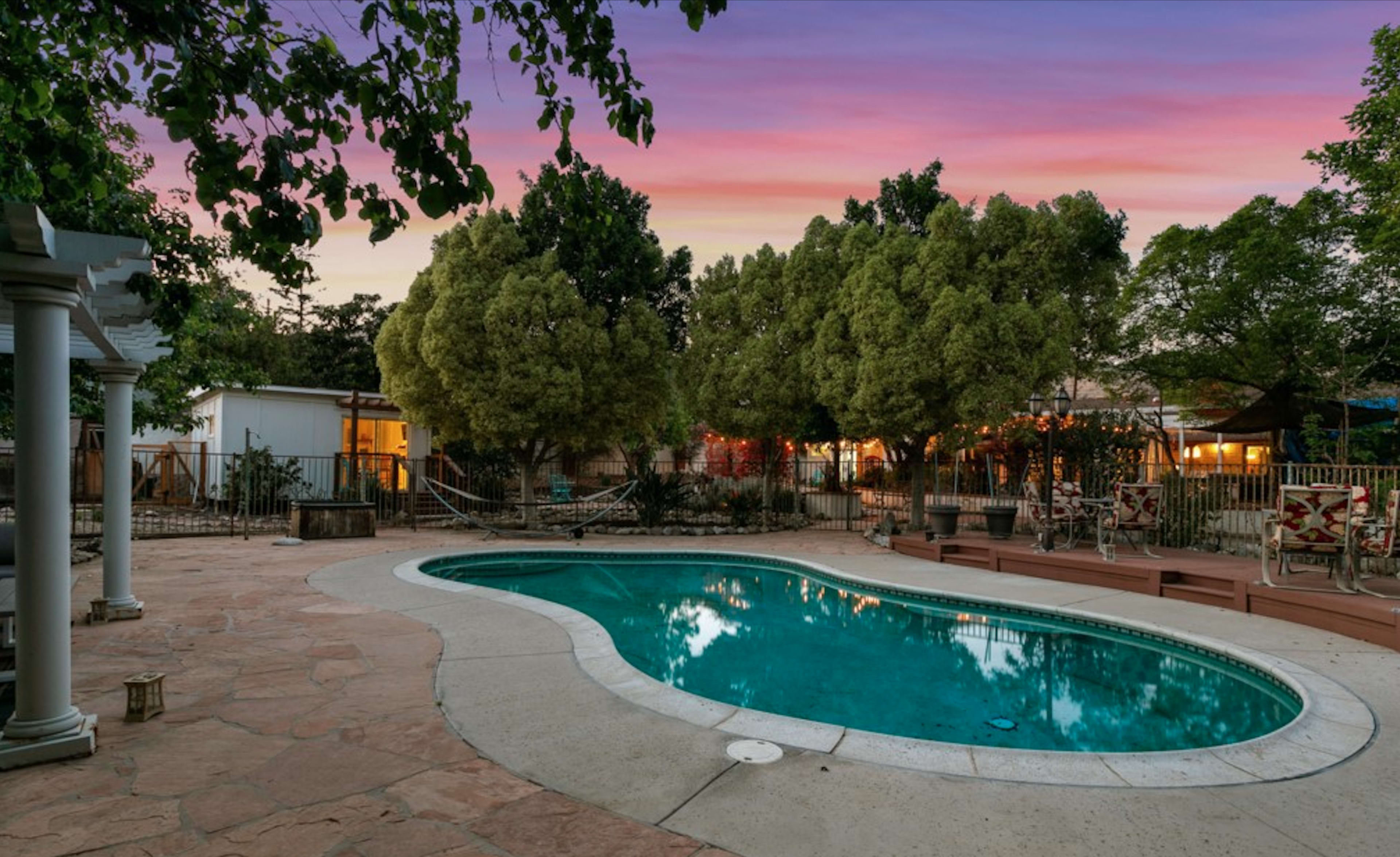 A swimming pool is surrounded by lush trees and patio furniture, with a sunset sky in the background.
