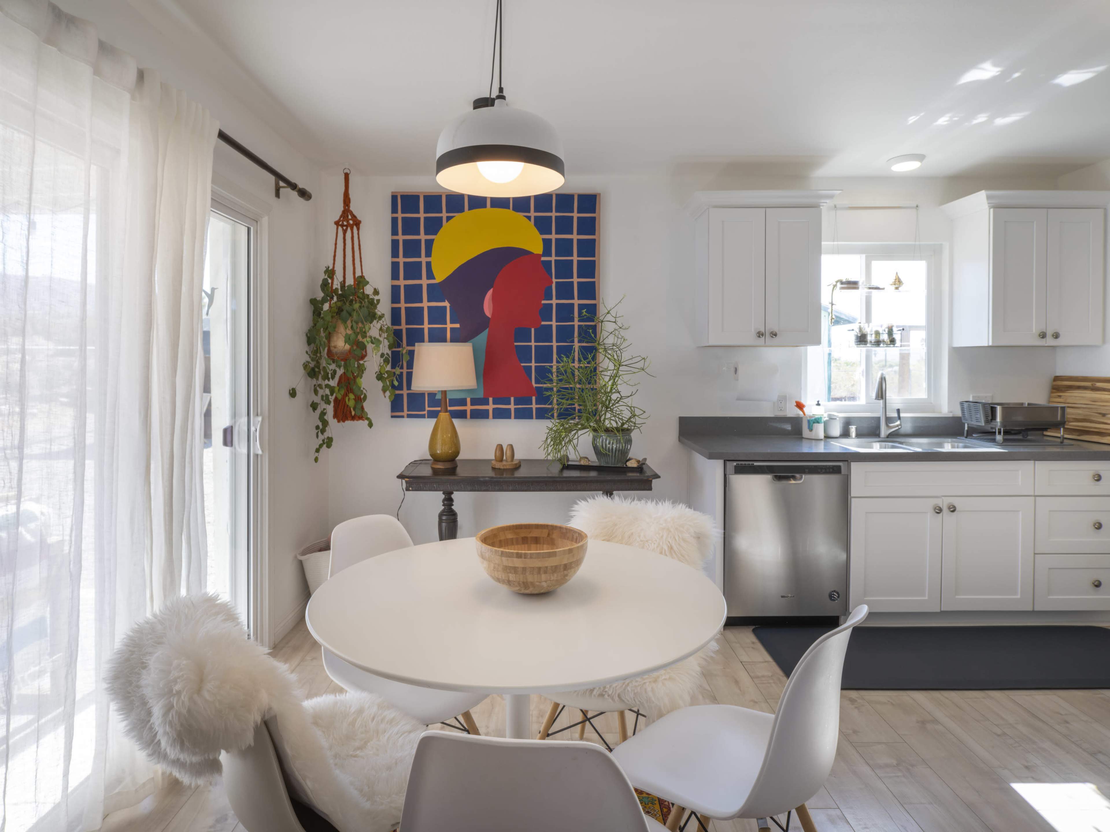 A bright kitchen features a round dining table with a wooden bowl, surrounded by white chairs, and is adorned with a colorful abstract wall art and plants.
