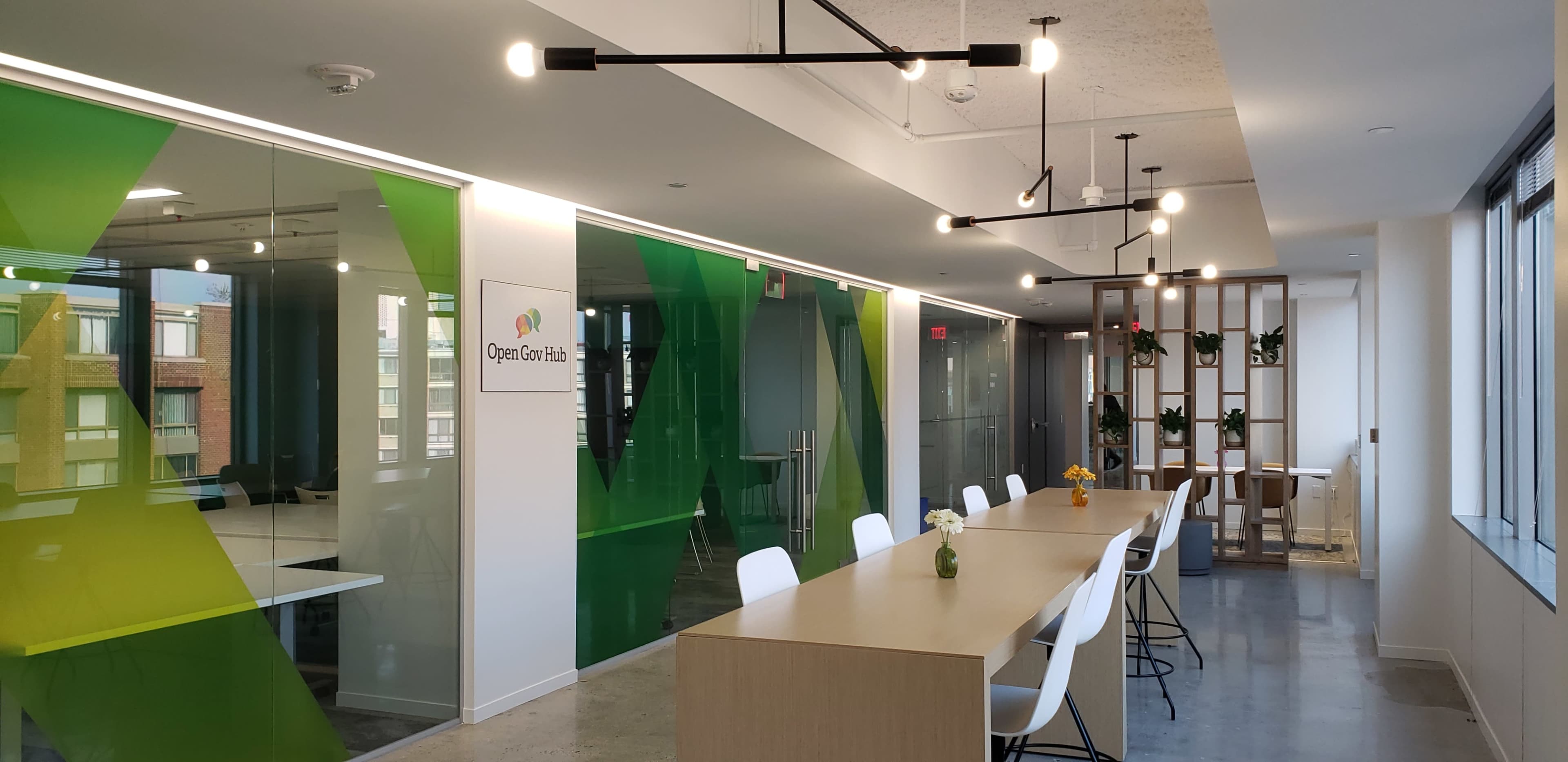 A modern office space featuring a long wooden table surrounded by white chairs, with large windows and green geometric glass partitions.