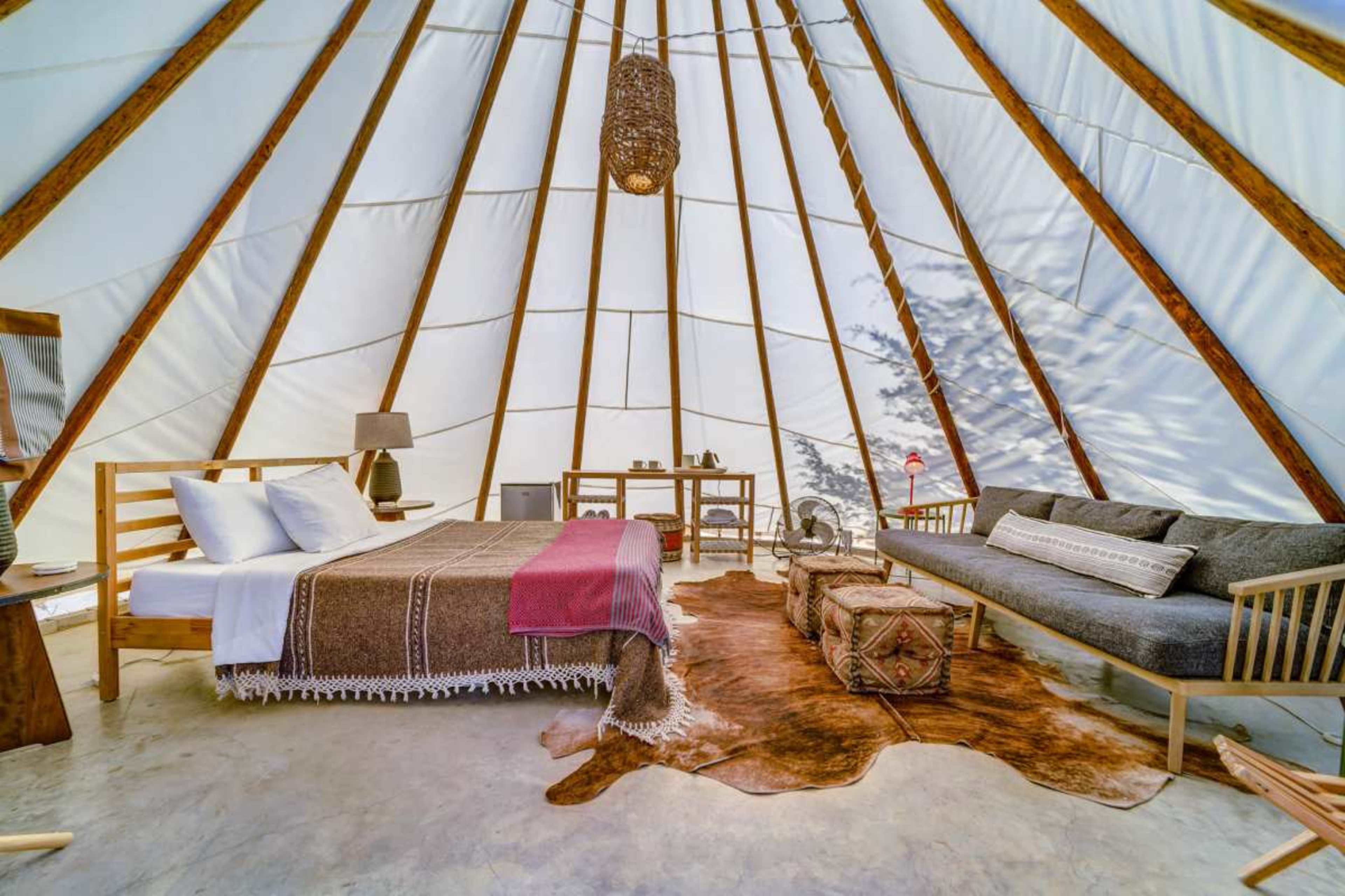 The interior of a glamping tent features a bed, a sofa, and natural wooden furnishings against a backdrop of white canvas walls.
