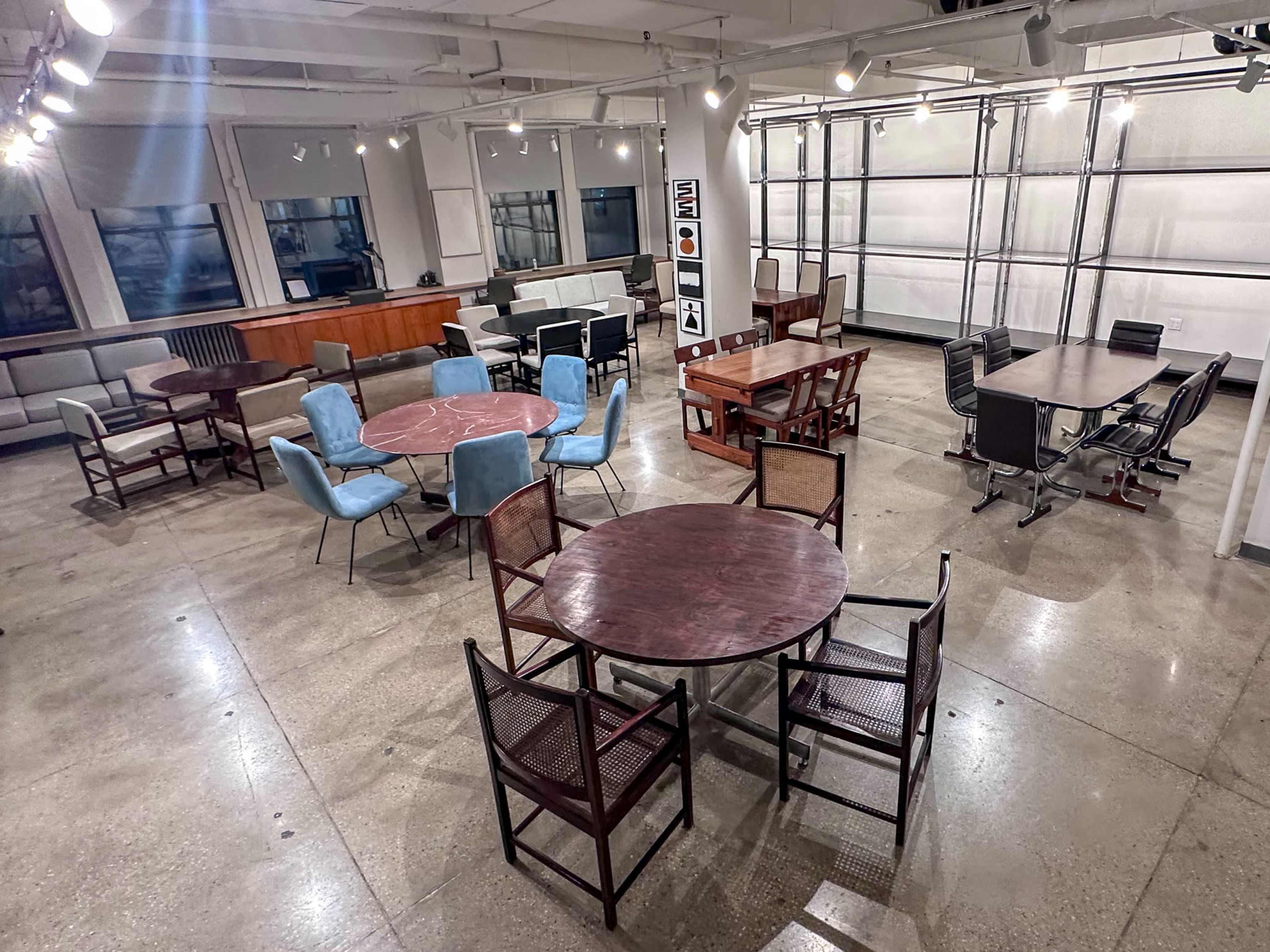 The image shows a spacious room with various tables and seating arrangements, featuring a mix of round and rectangular tables surrounded by different styles of chairs.