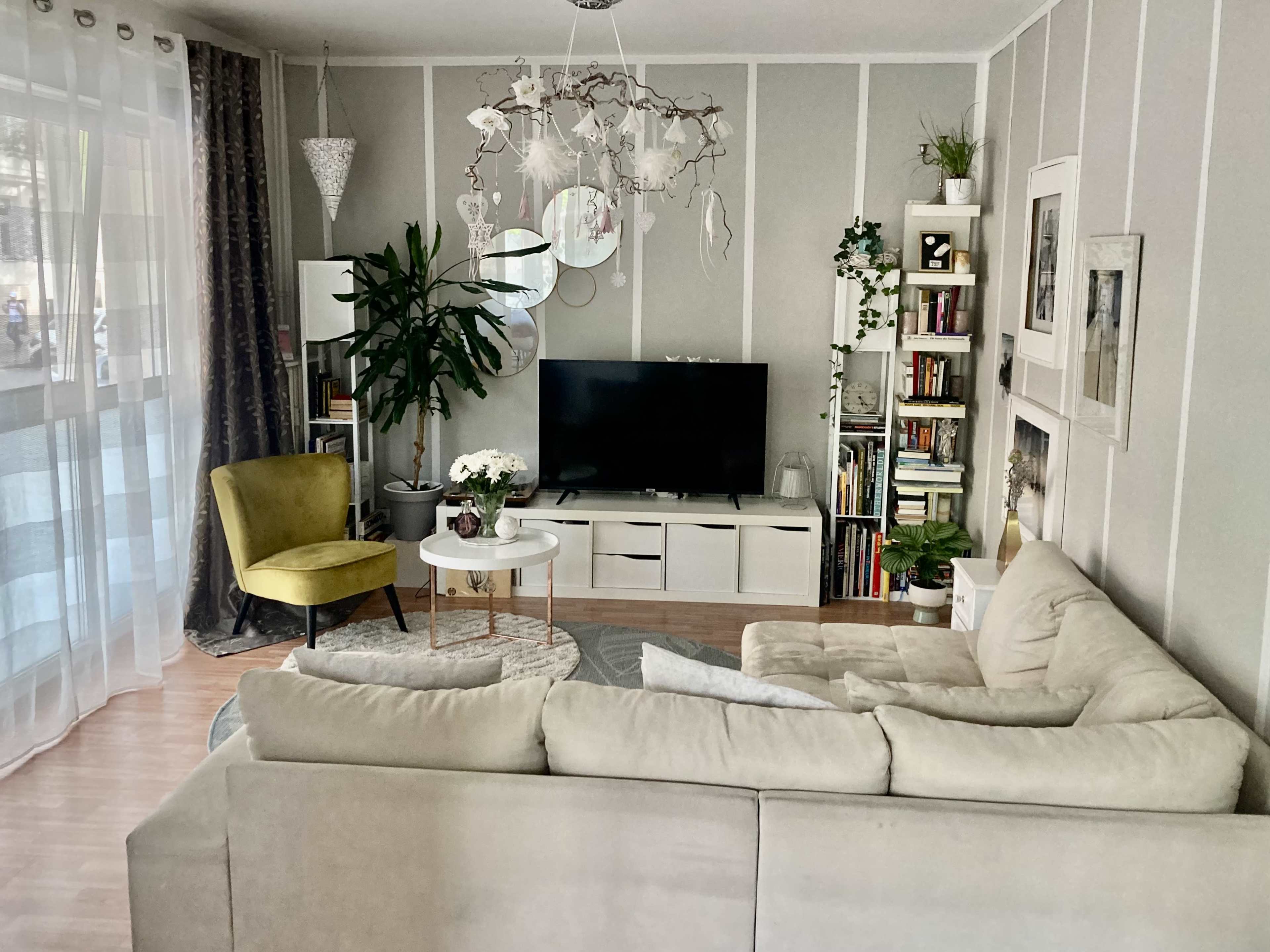 The image shows a living room with a beige sectional sofa, a green accent chair, a television on a white entertainment unit, and plants decorating the space.