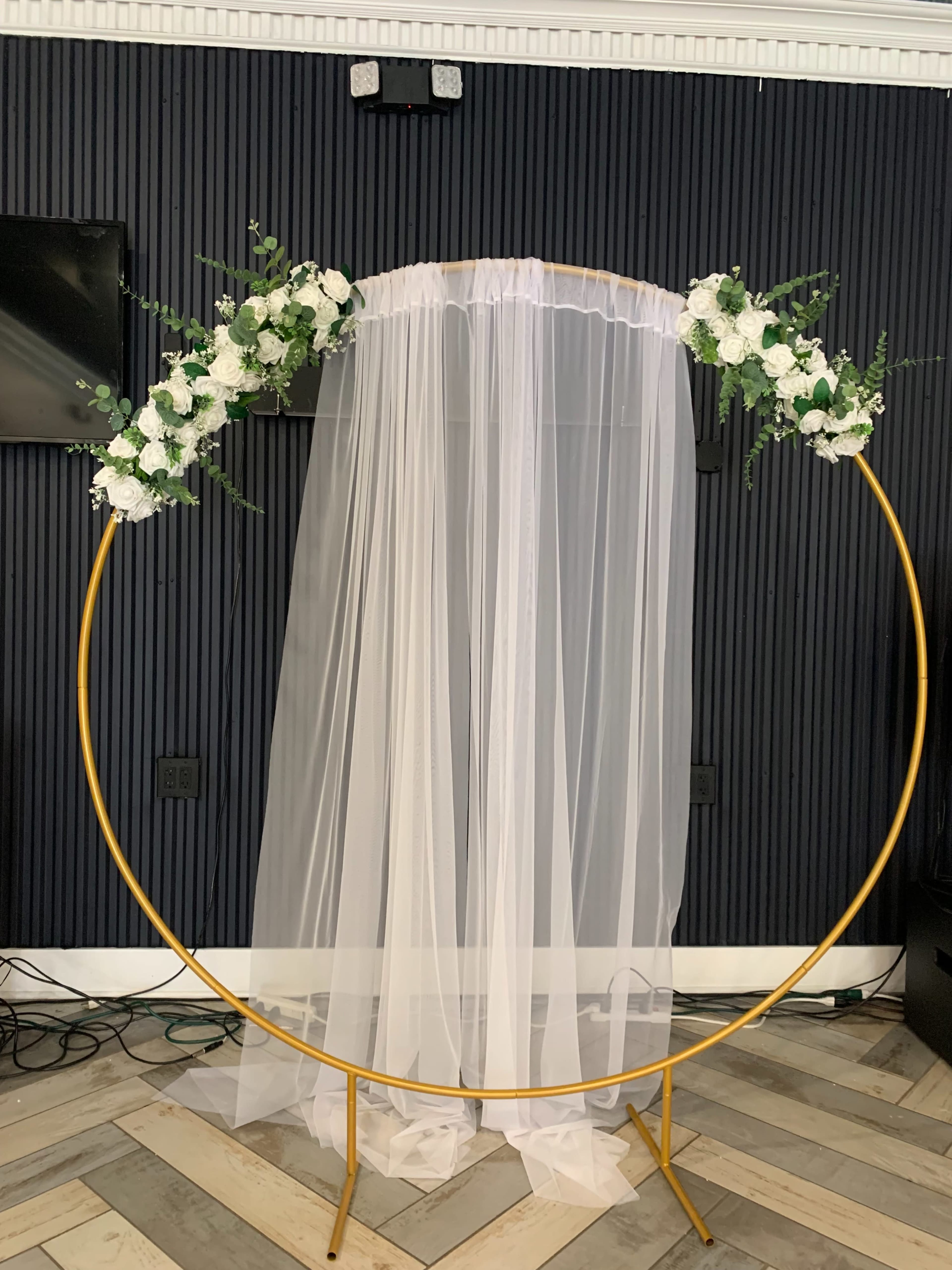 A circular backdrop stand adorned with white flowers and a sheer white curtain, set against a dark wall.