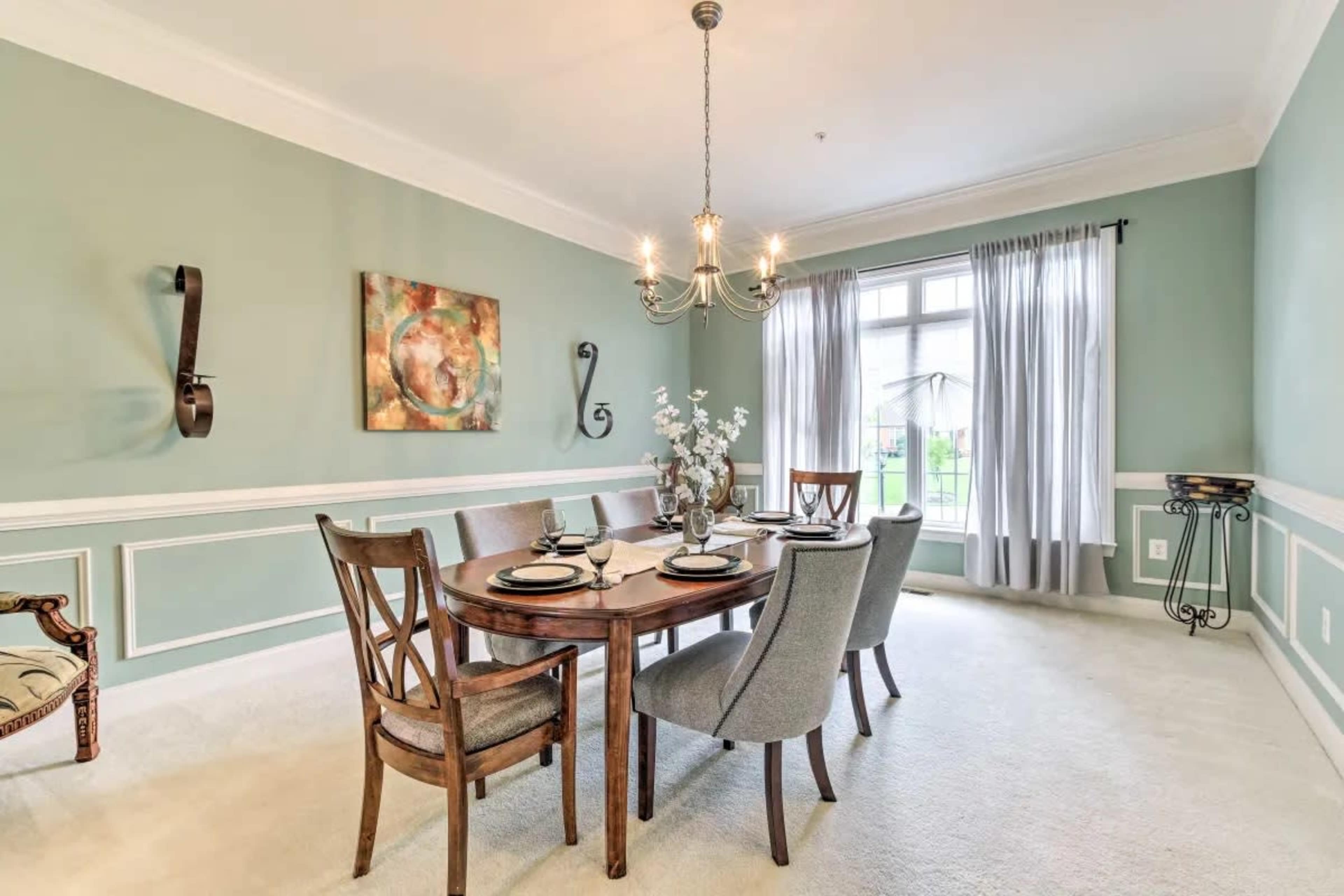 A dining room features a round table set with dishes, surrounded by wooden and upholstered chairs, under a chandelier, with light green walls and a window that lets in natural light.