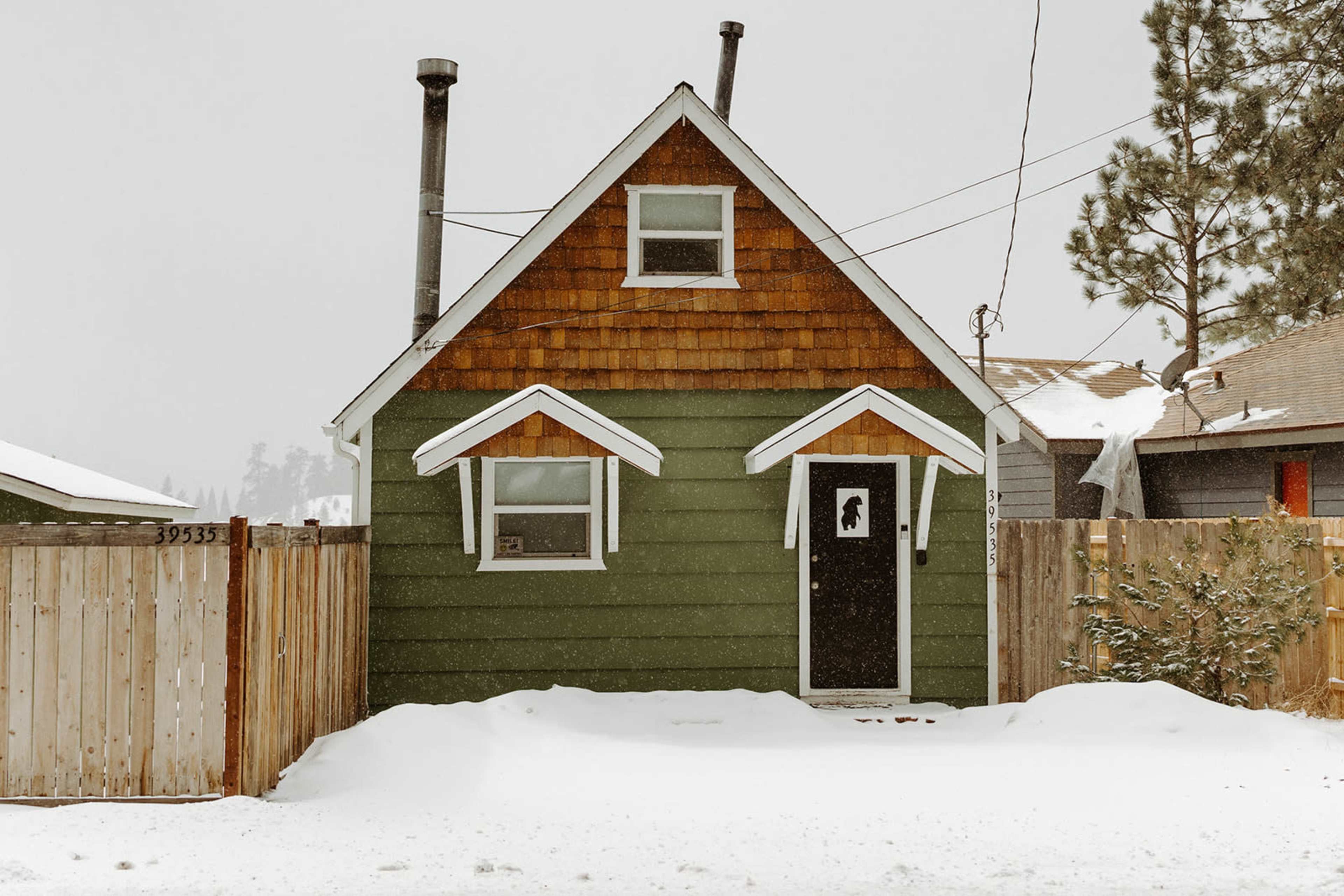 A green house with a brown-shingled roof and white accents is partially buried in snow, set against a snowy backdrop and surrounded by a wooden fence.