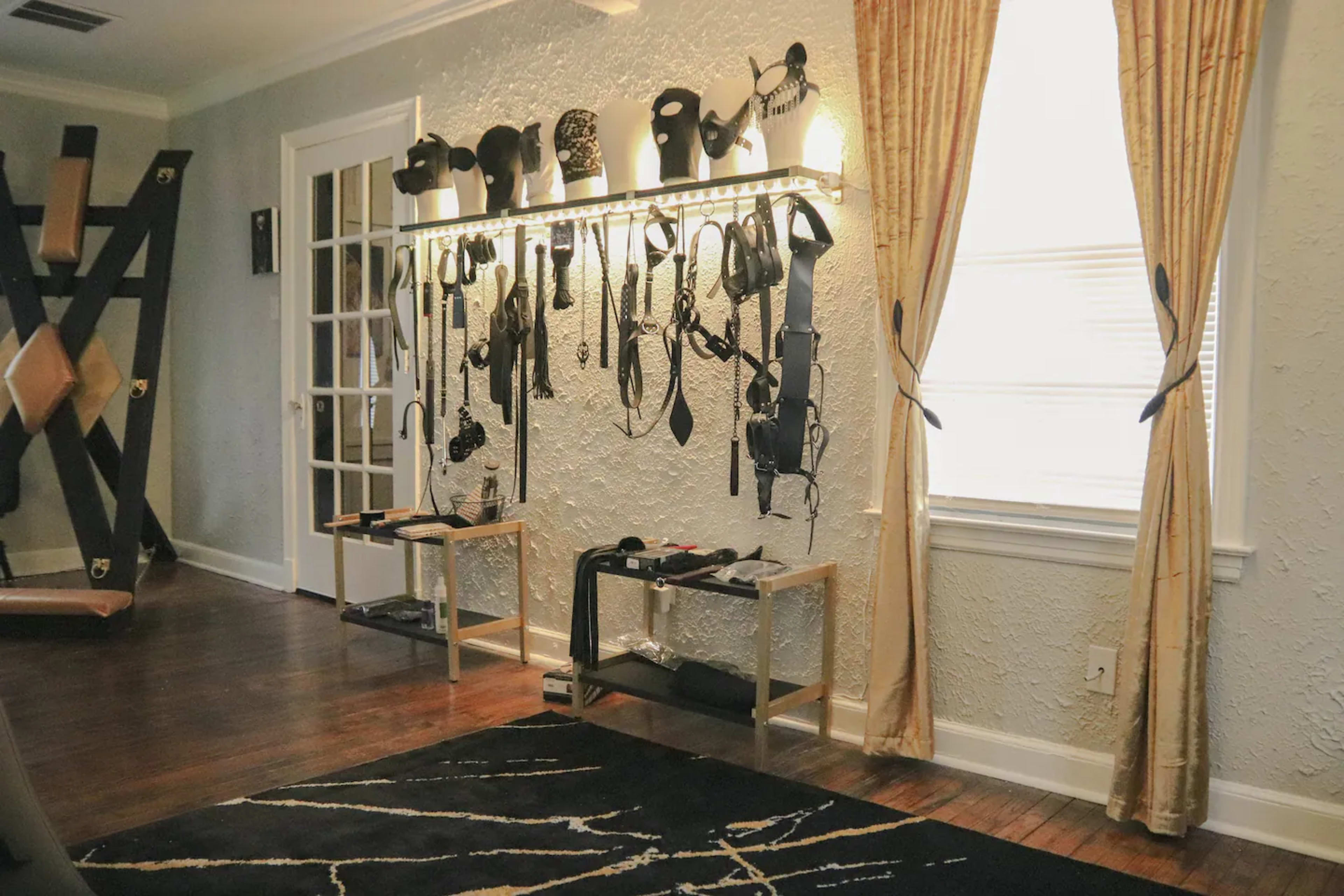 The room features a wall-mounted display with various harnesses and equipment, complemented by a curtain-draped window and wooden flooring.