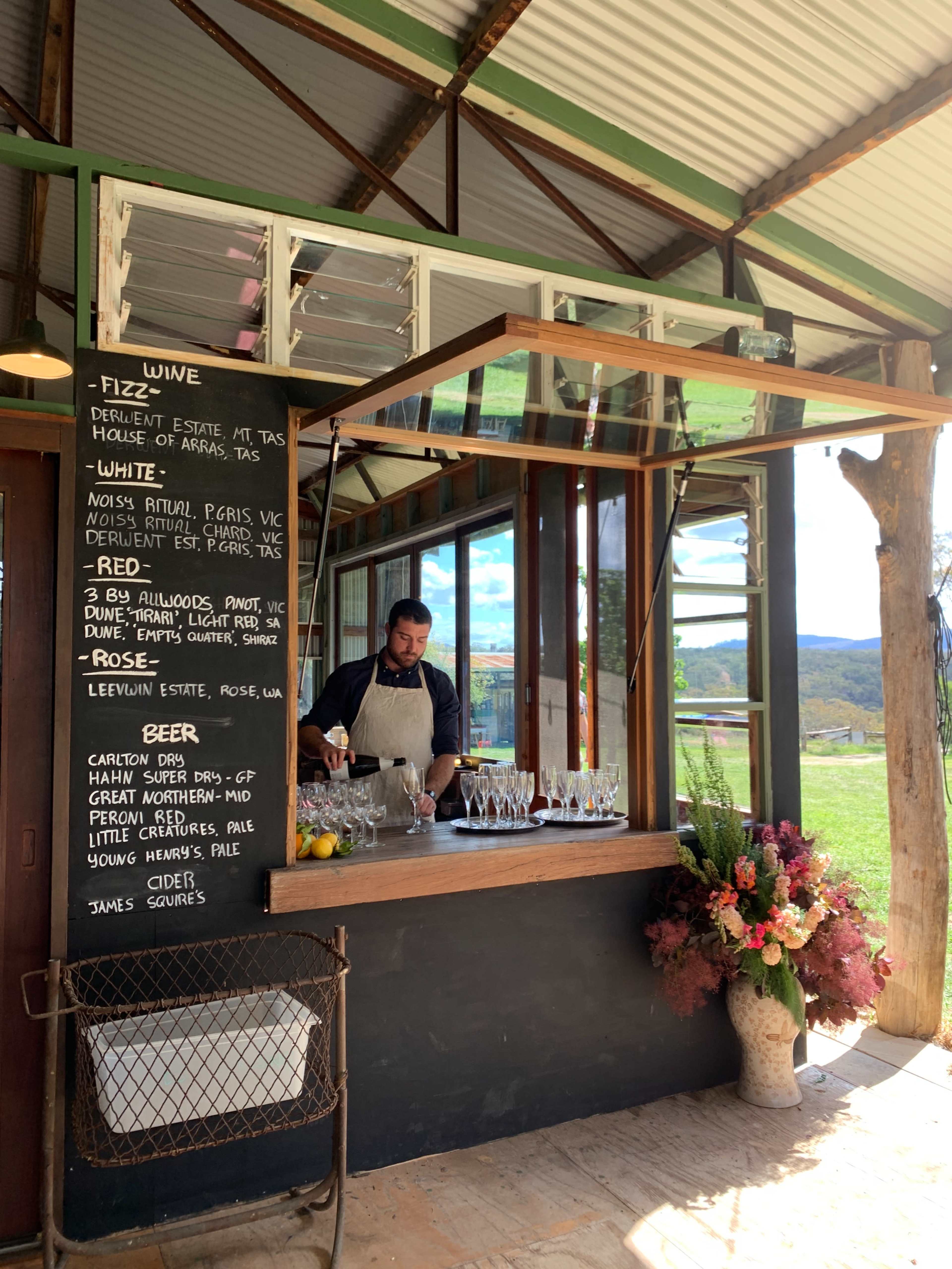 A man in an apron stands behind a wooden bar serving wine in a rustic setting with large windows and floral decorations.