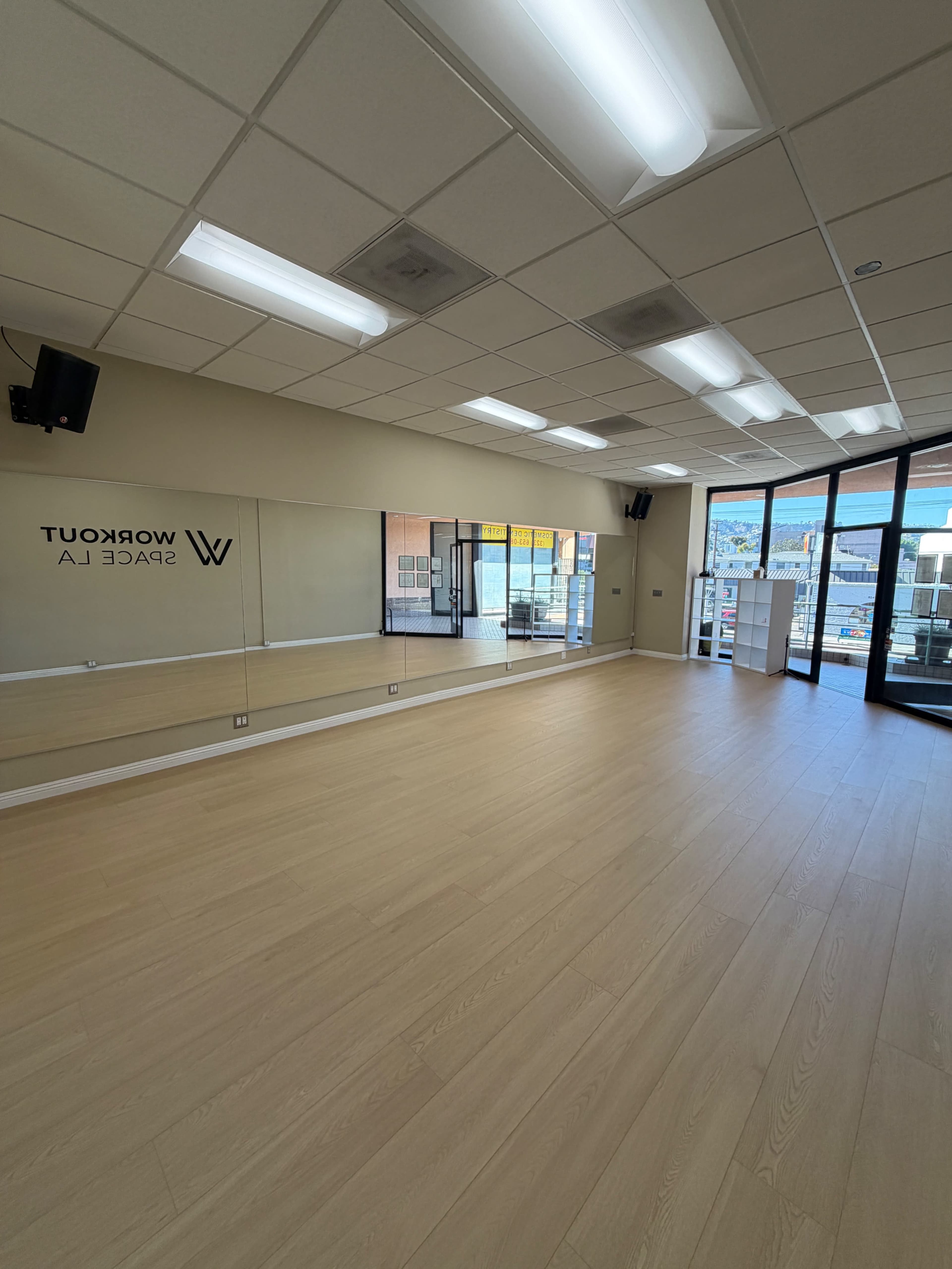 The image shows a spacious room with wooden flooring, large windows, and a mirrored wall, designed for workouts or exercise classes.