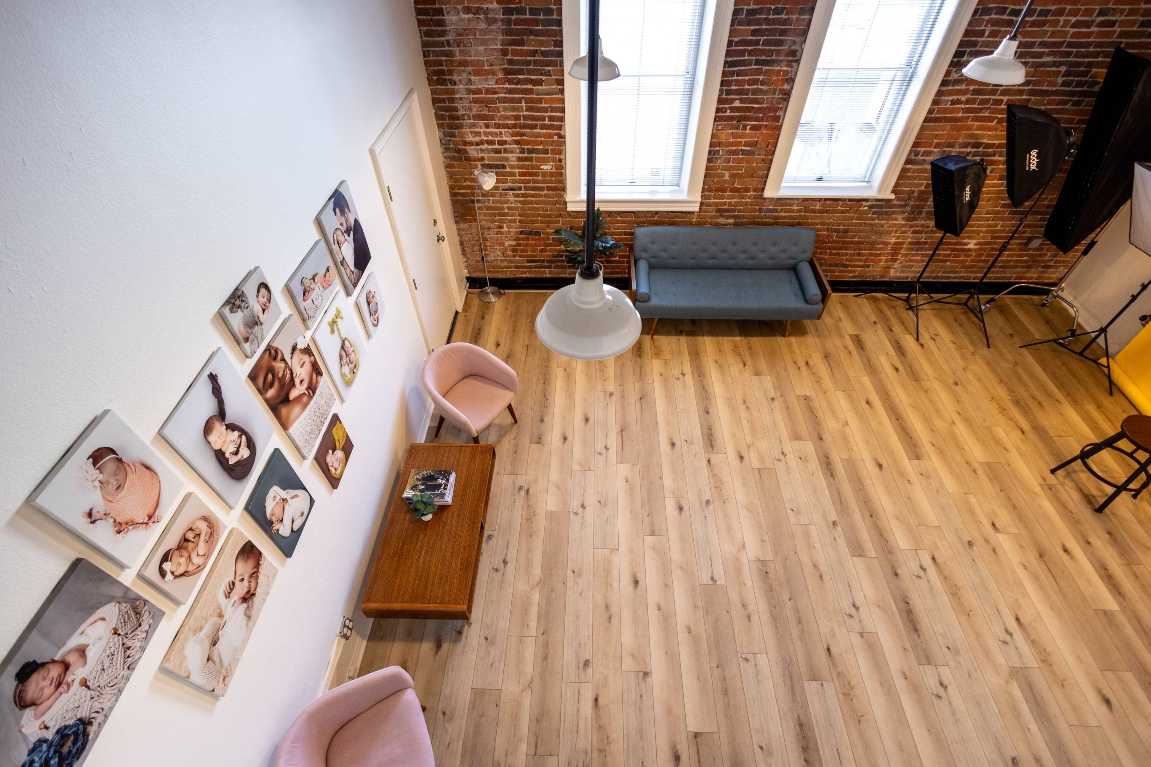 The image shows a spacious interior featuring hardwood flooring, large windows, a gray sofa, two pink chairs, a wood table, and several framed photographs on the wall.