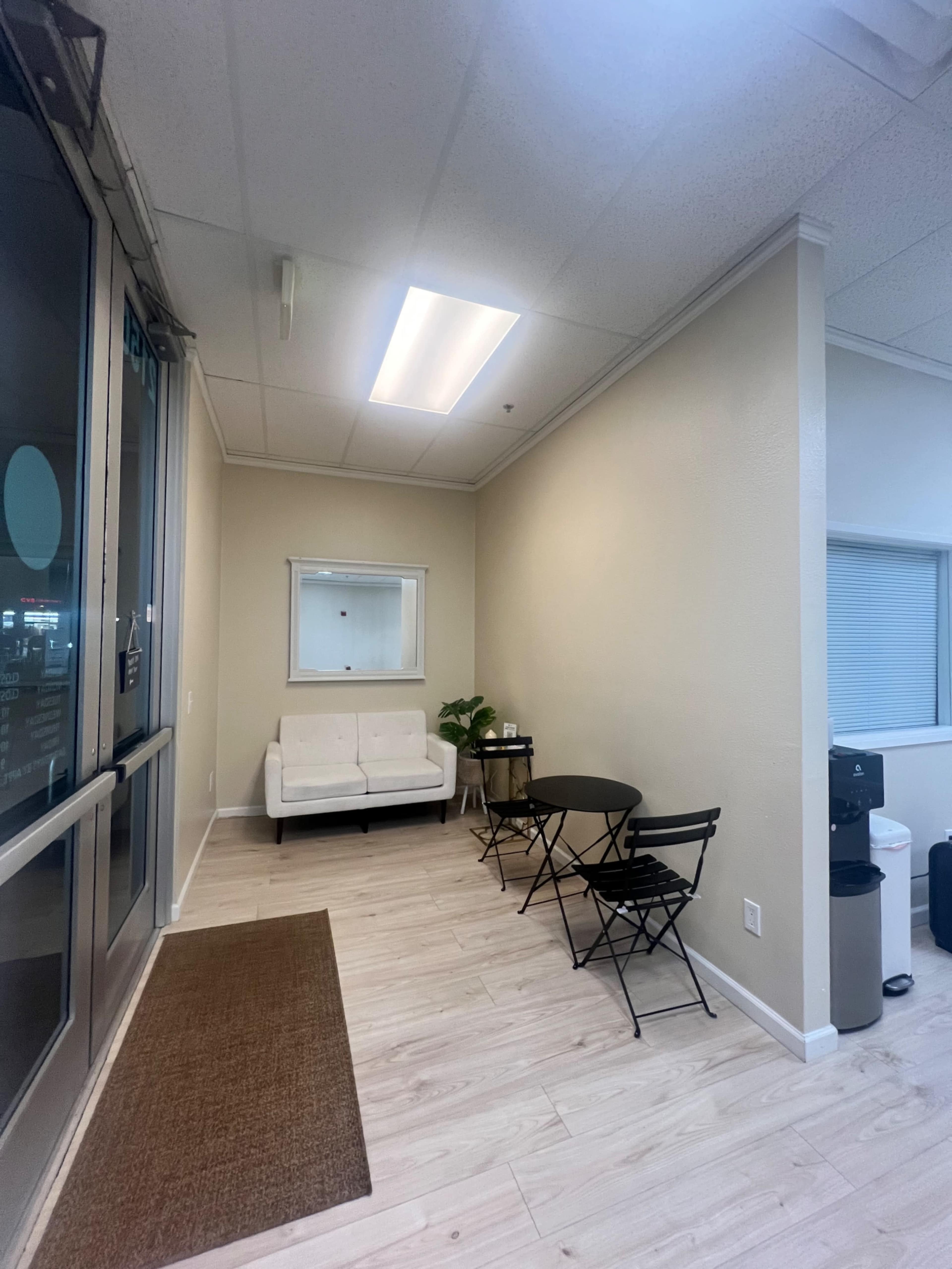 A small waiting area with a white sofa, a round table, and two chairs, all situated against a light-colored wall.