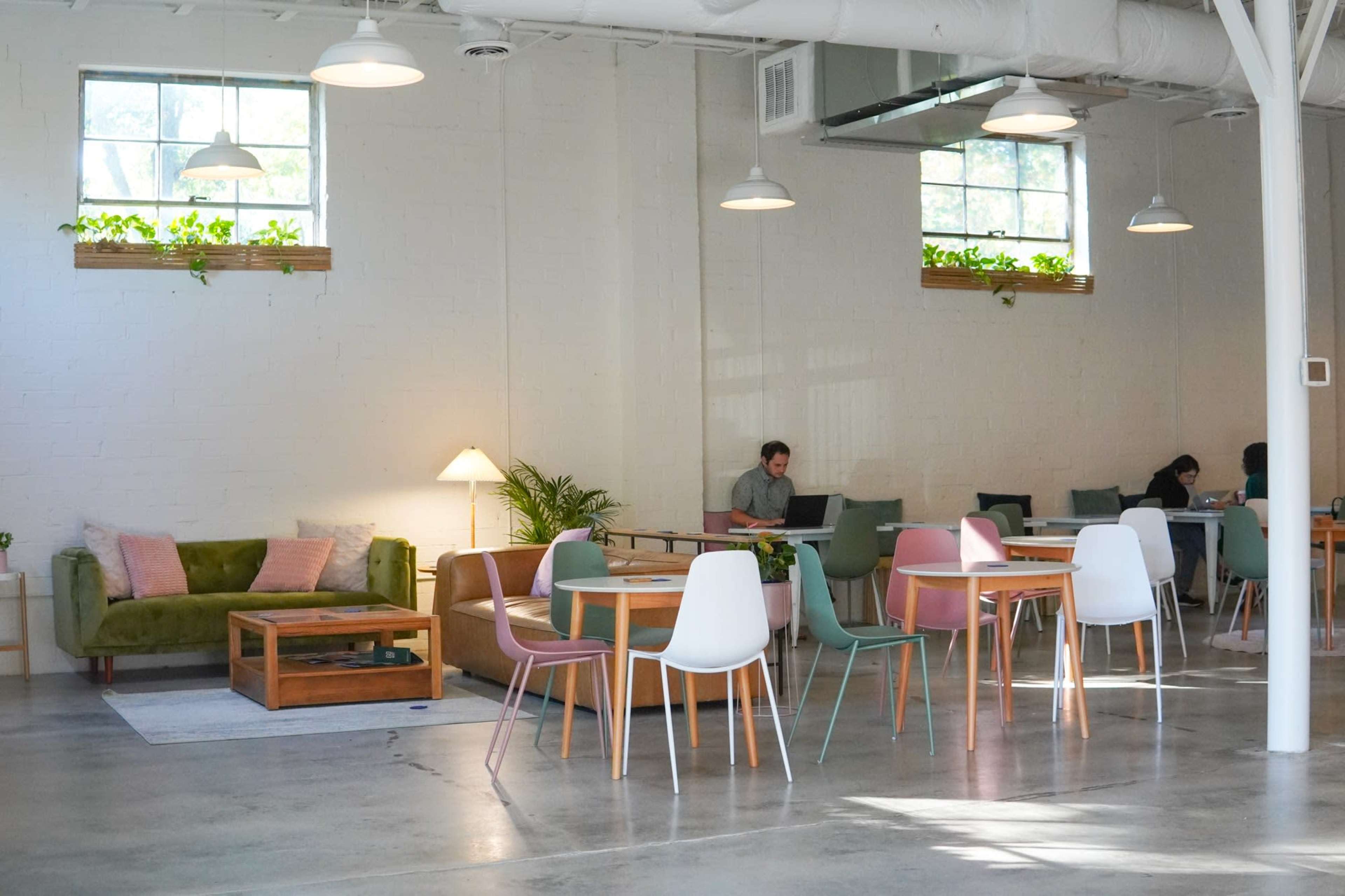 The image shows a bright, airy co-working space featuring various tables, chairs, and a green sofa, with natural light filtering through large windows.