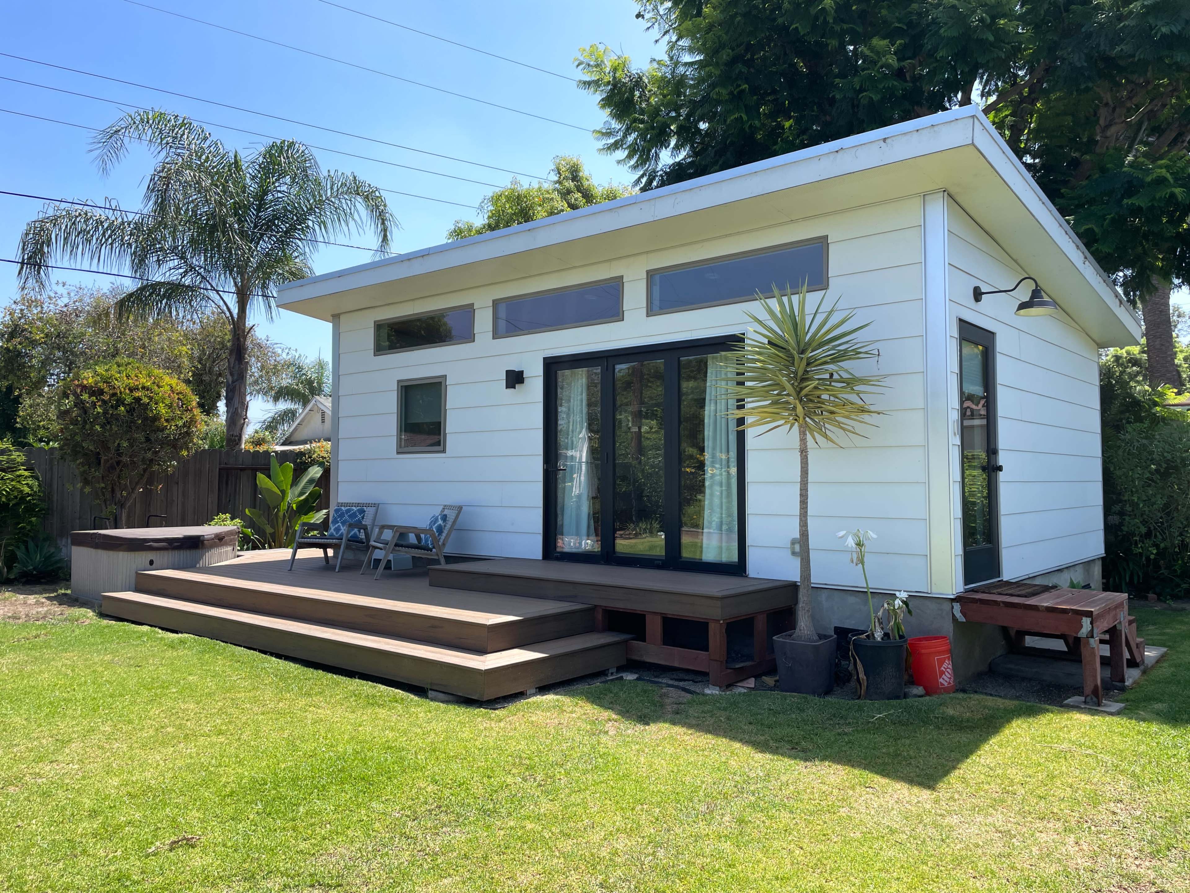 A small, modern white house features a wooden deck, large windows, and palm trees in the surrounding yard.