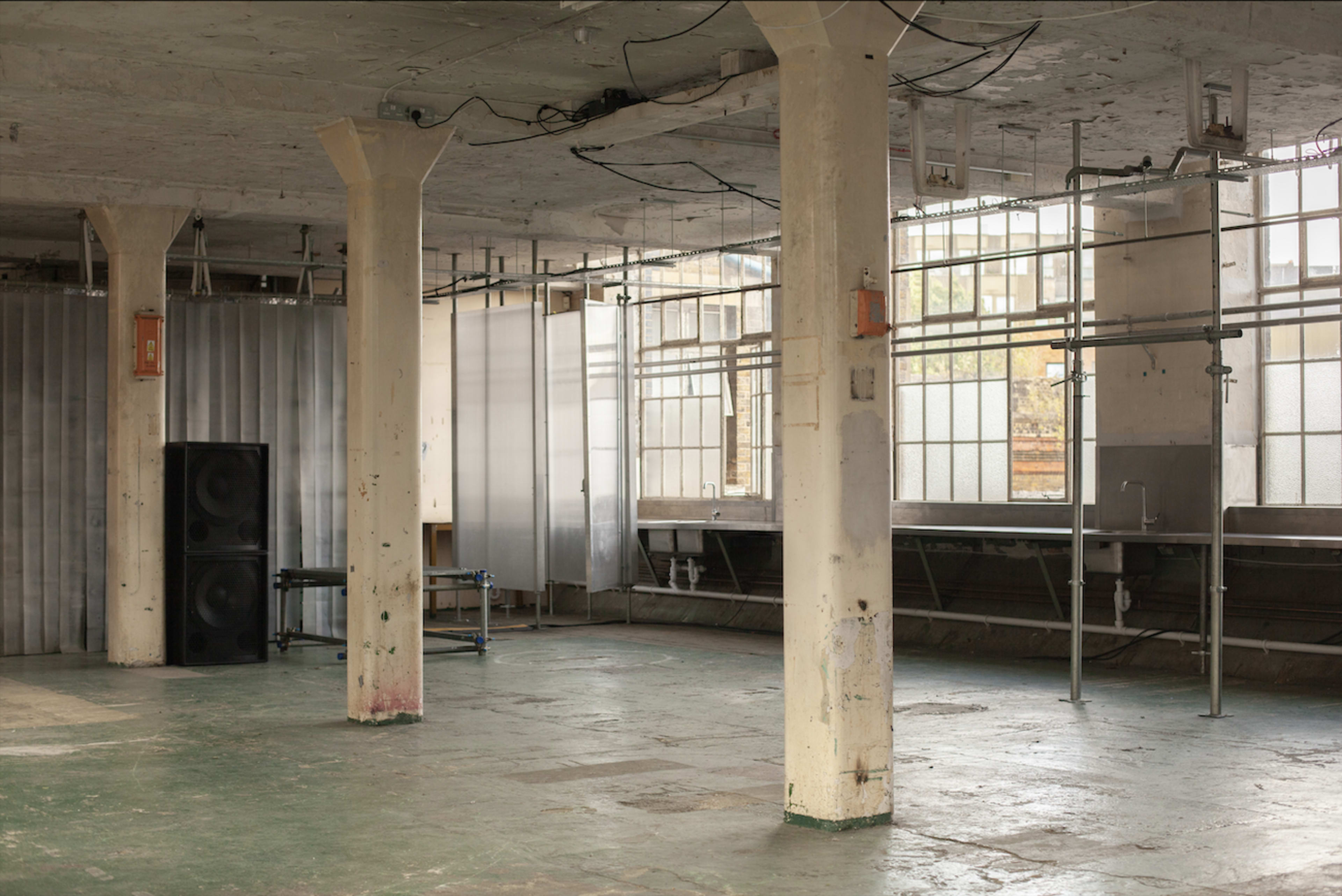 The image shows an empty, industrial space with concrete pillars, exposed wiring, and large windows, featuring scaffolding and a pair of speakers in one corner.