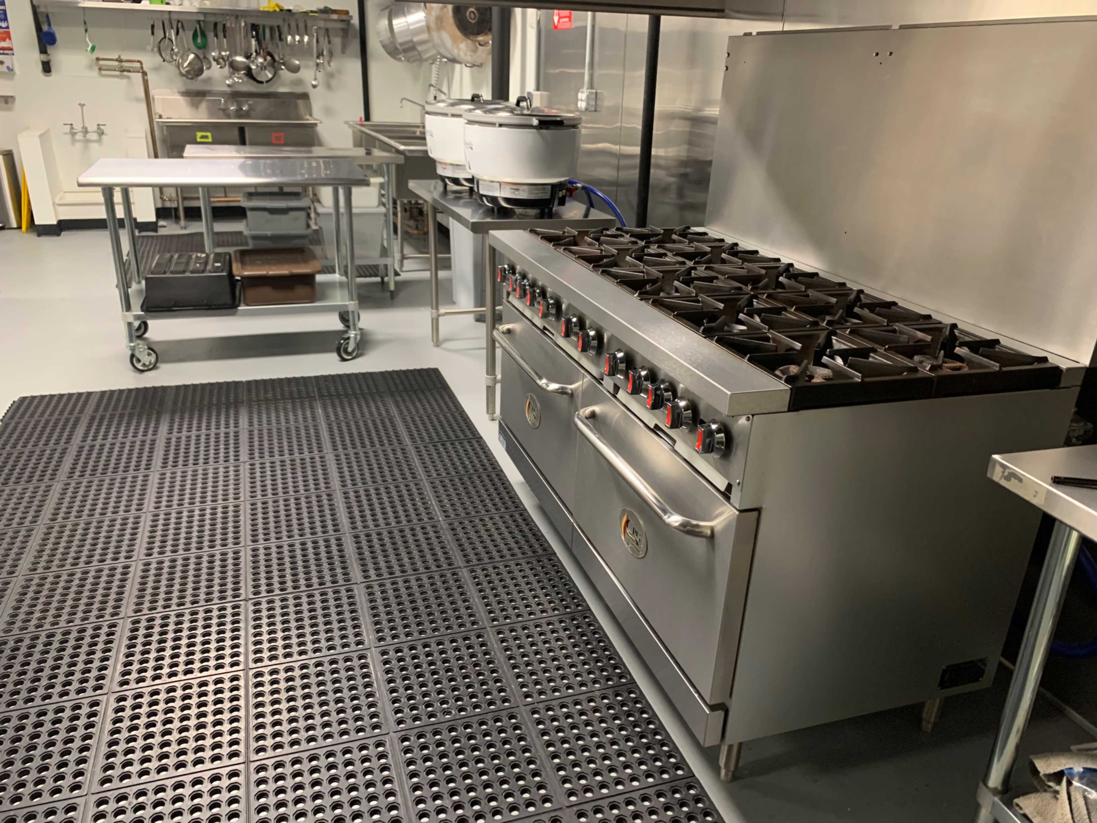 A stainless steel commercial kitchen with a large gas range, a metal prep table, and a textured black mat on the floor.