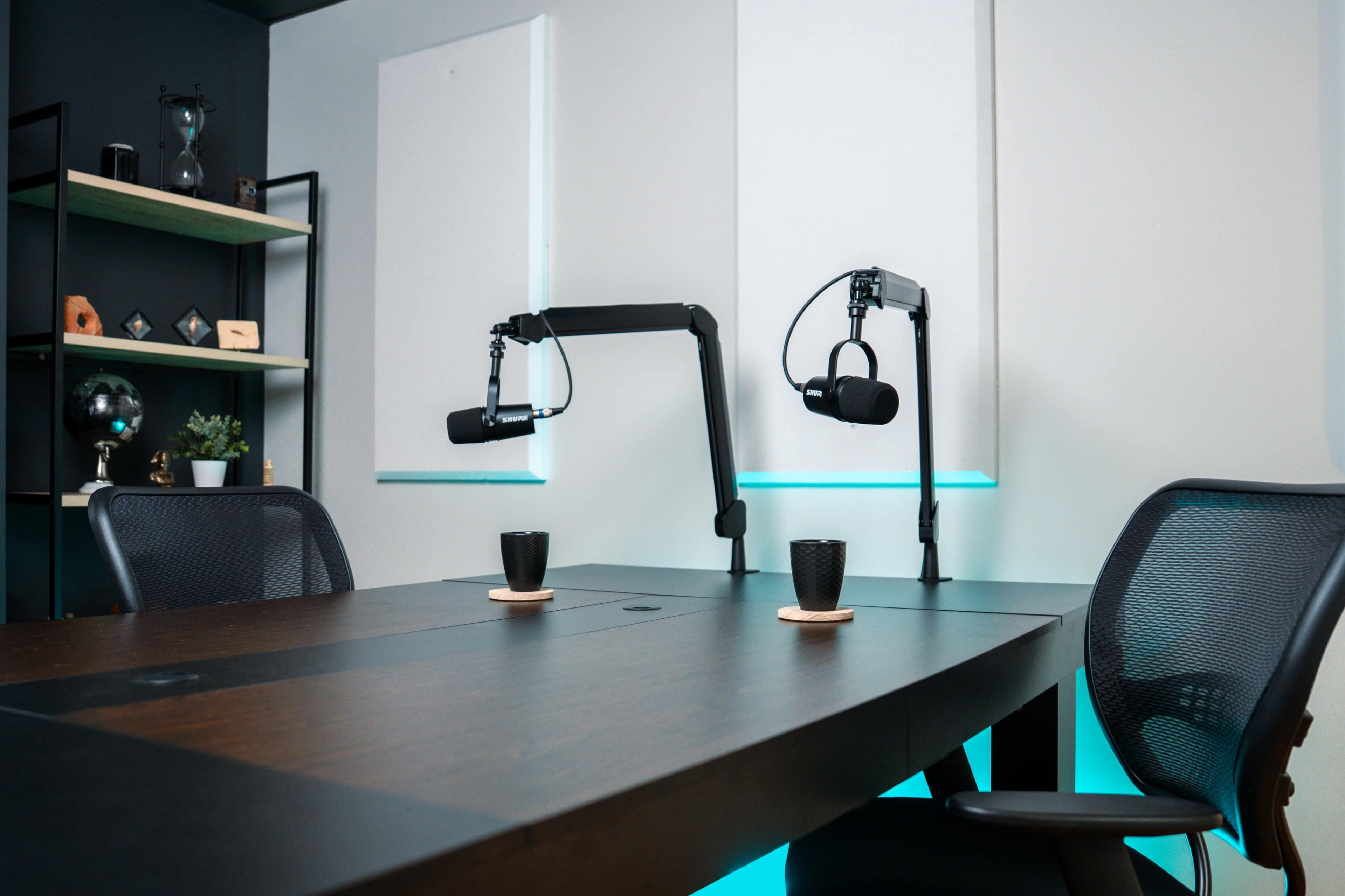 The image shows a modern podcasting studio featuring a large table, two microphones mounted on stands, and two black cups on coasters.