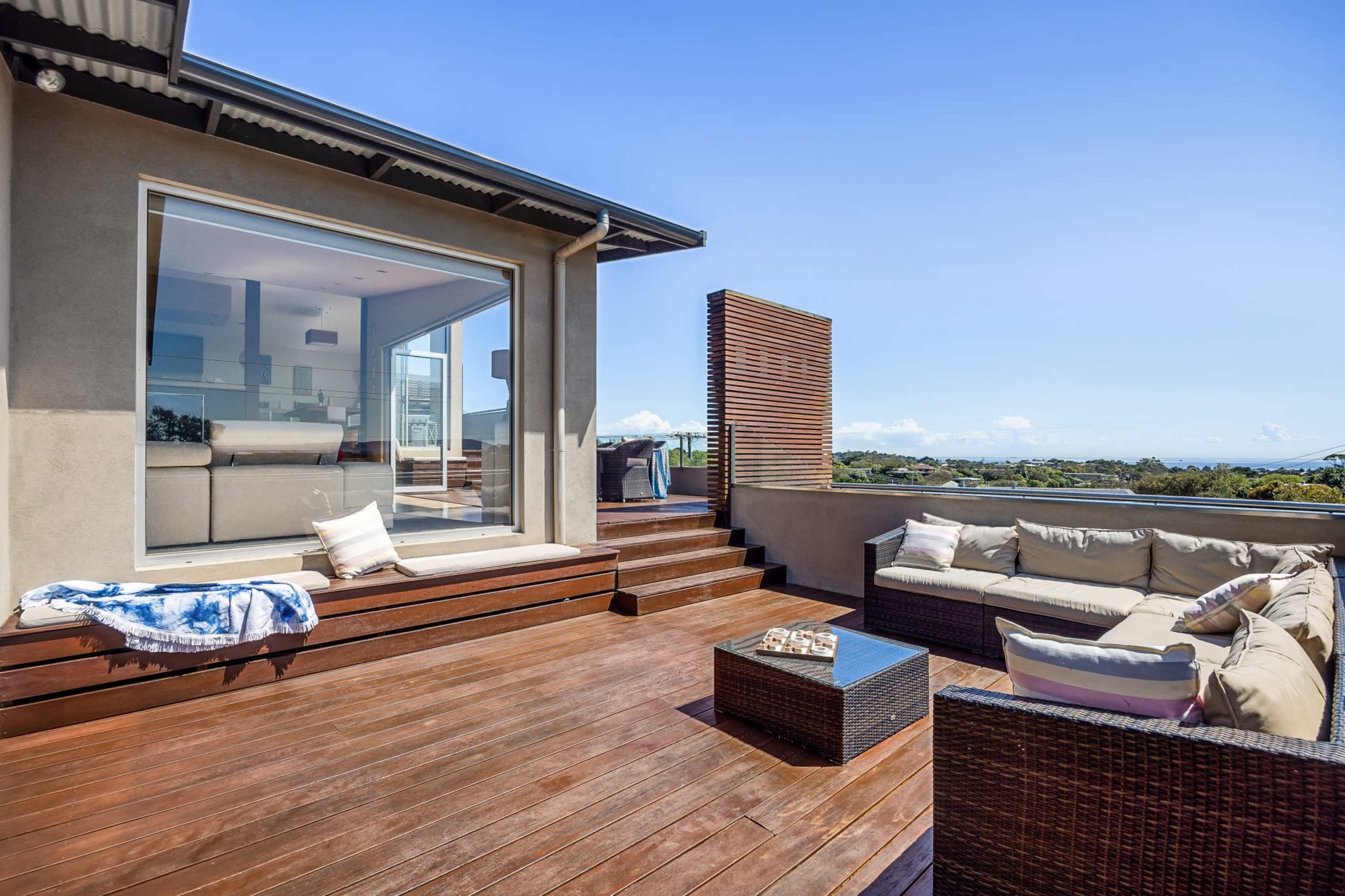 A modern patio features a seating area with cushions, a glass table, and wooden flooring, overlooking a scenic view.