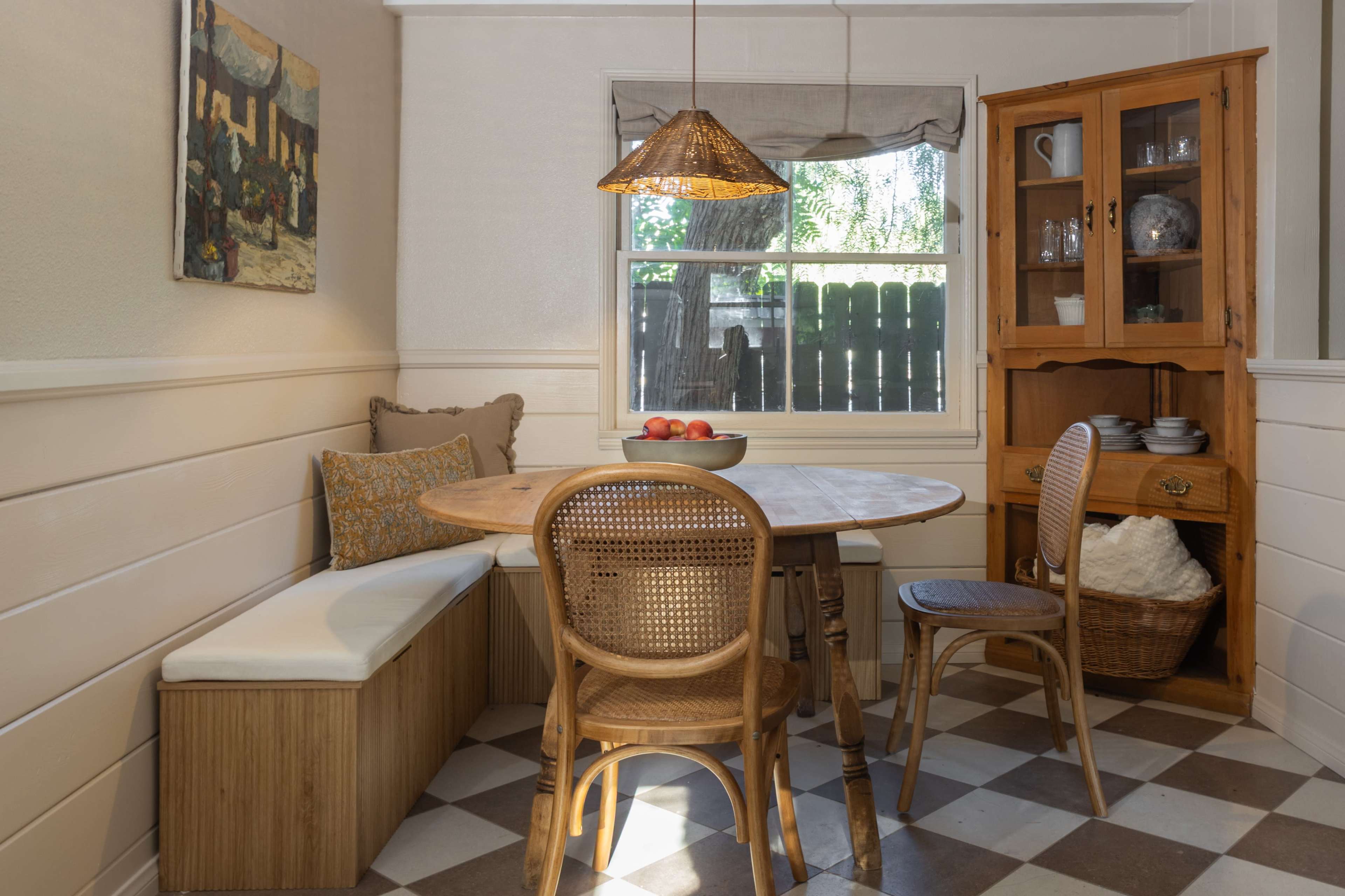 A cozy breakfast nook features a round wooden table surrounded by chairs, a built-in bench, and a cabinet with dishes, all under a pendant light.