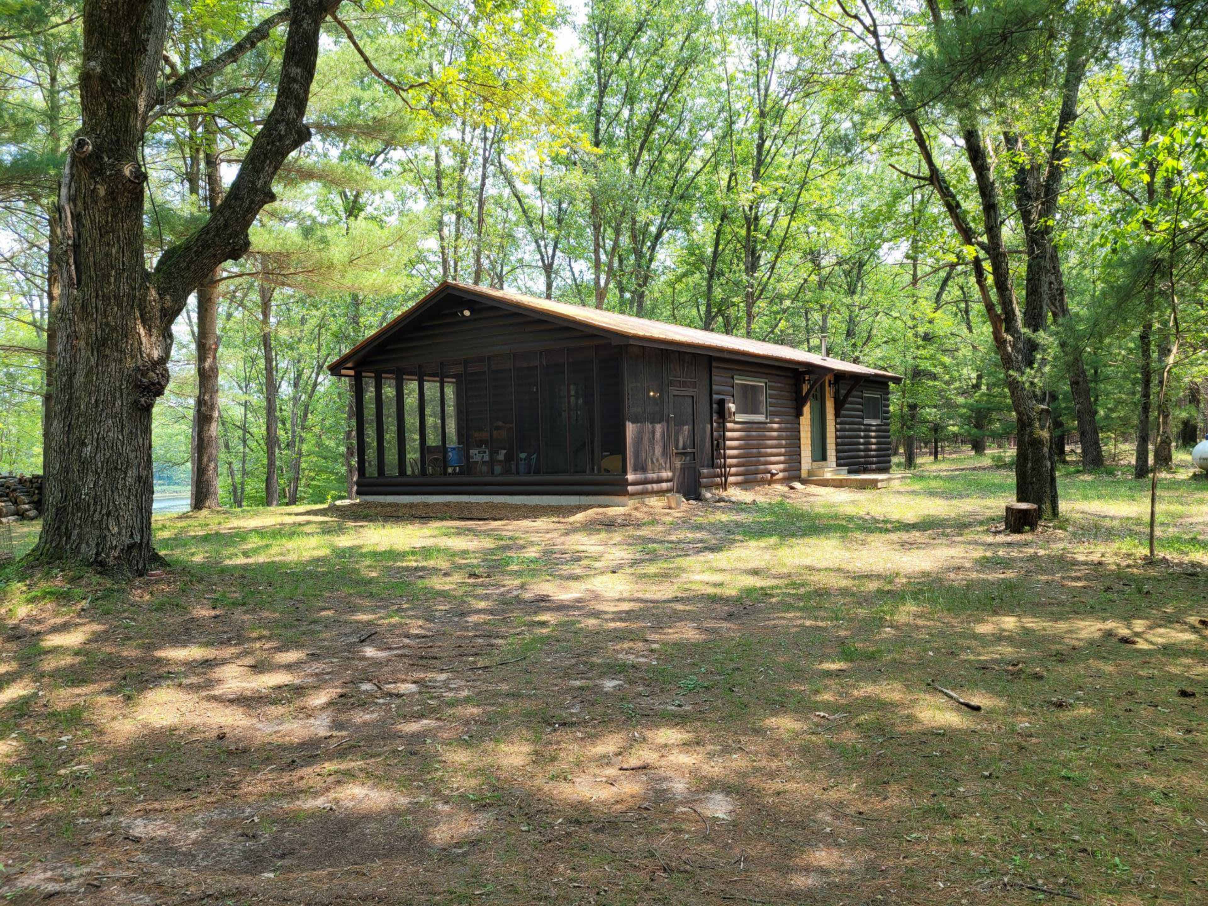 A wooden cabin is situated among trees, featuring a screened porch and a clear view of the surrounding forest.