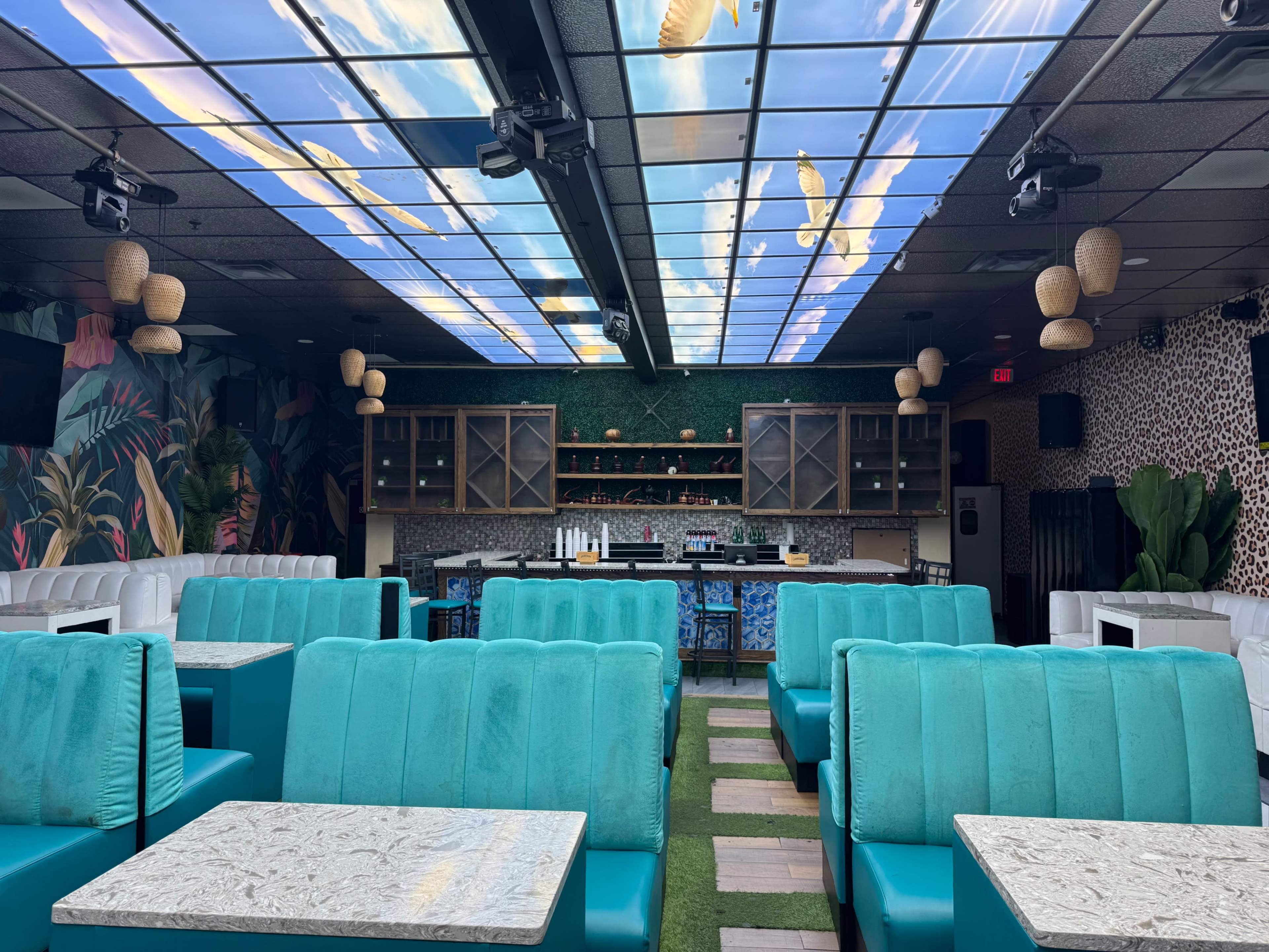 The image shows a brightly decorated bar area with turquoise seating, a lush green ceiling design, and a well-stocked bar in the background.