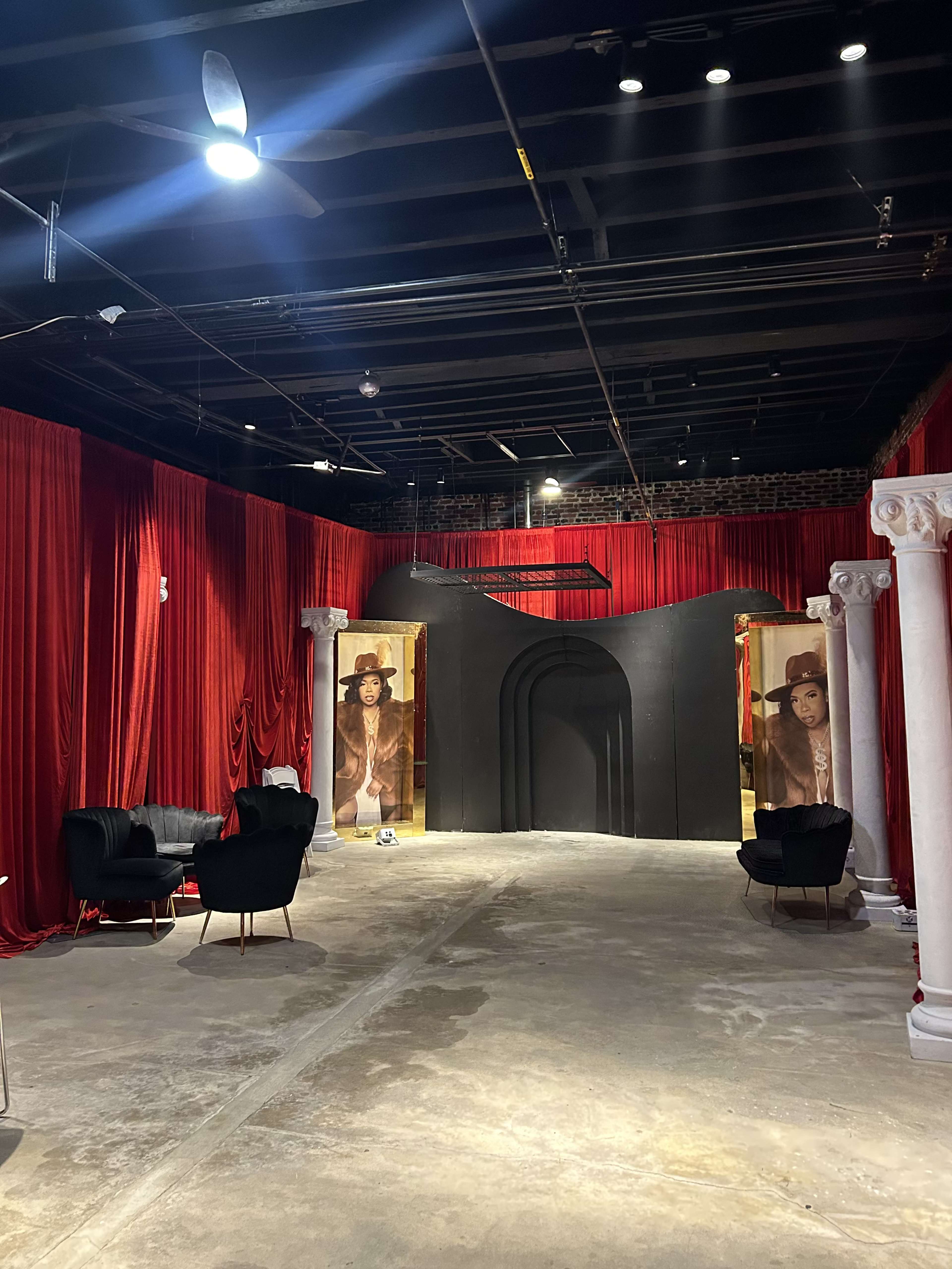The image shows an interior space with red drapery, two large black doorways flanked by columns, and a pair of black chairs arranged in the center.
