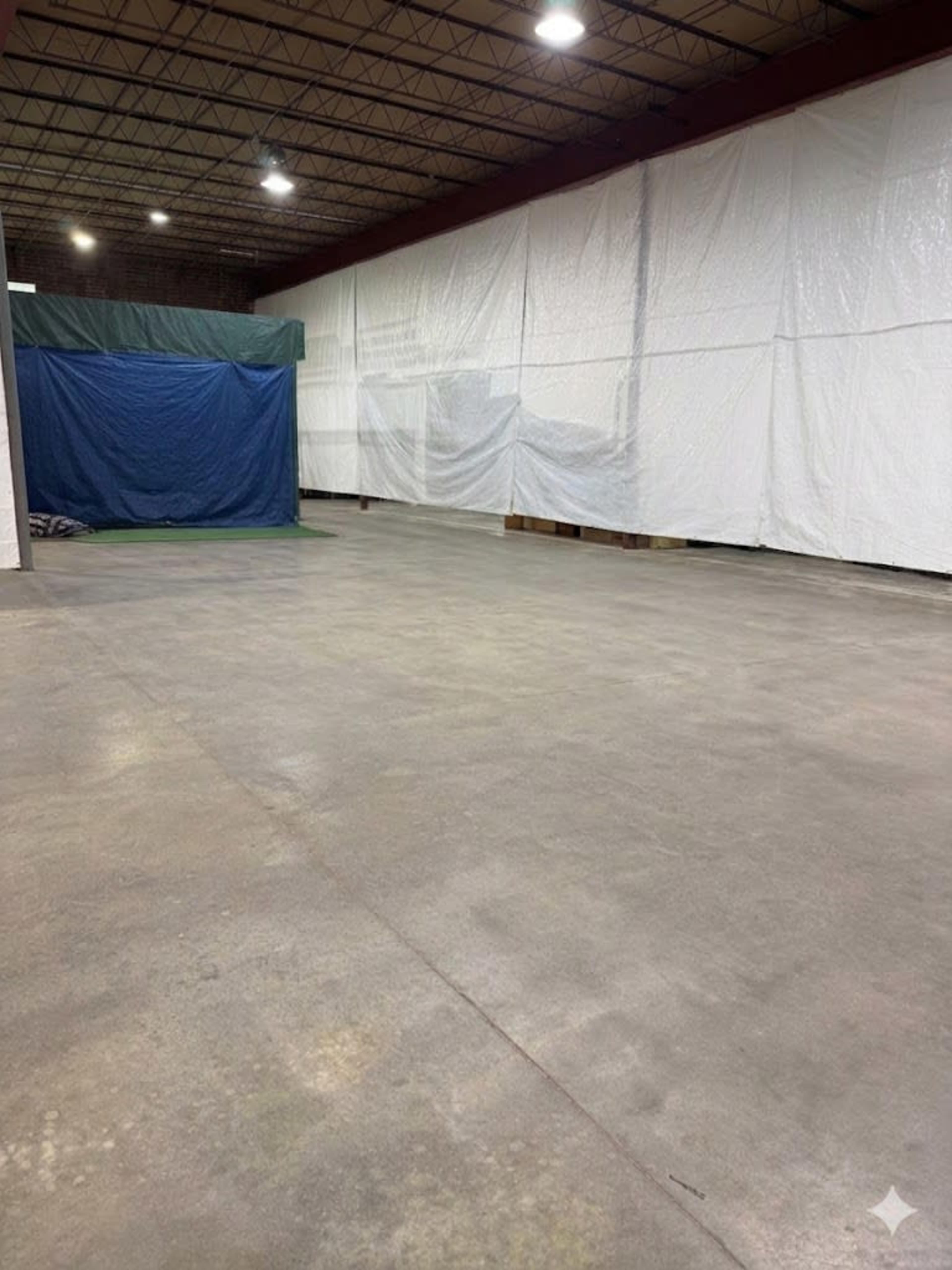The image shows an expansive, empty indoor space with concrete flooring, partially divided by white tarps and a blue tarp at the far end.