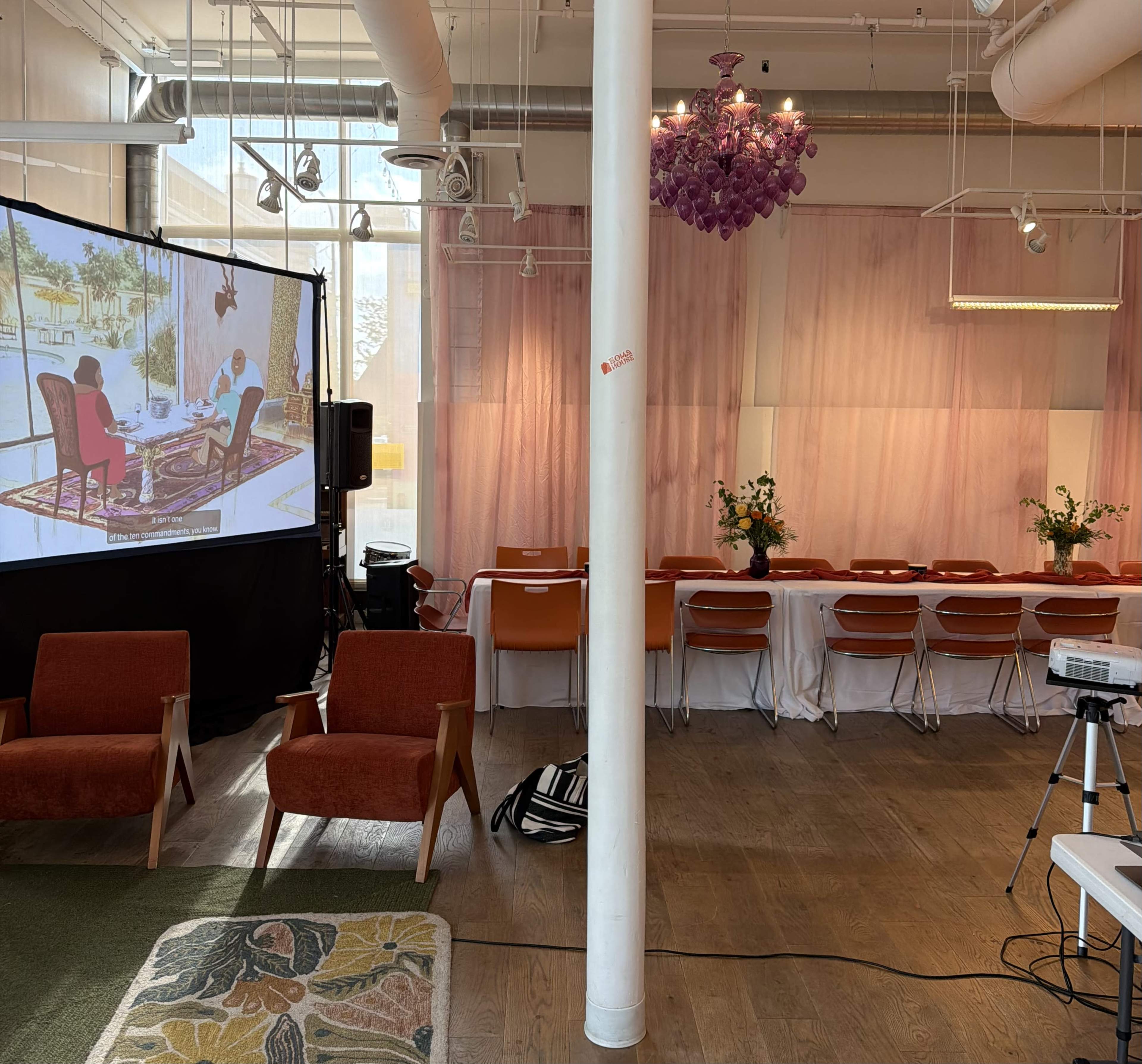 The image shows a spacious room with a presentation screen displaying a scene, two orange armchairs, a long table set with flowers, and a booth with a projector.