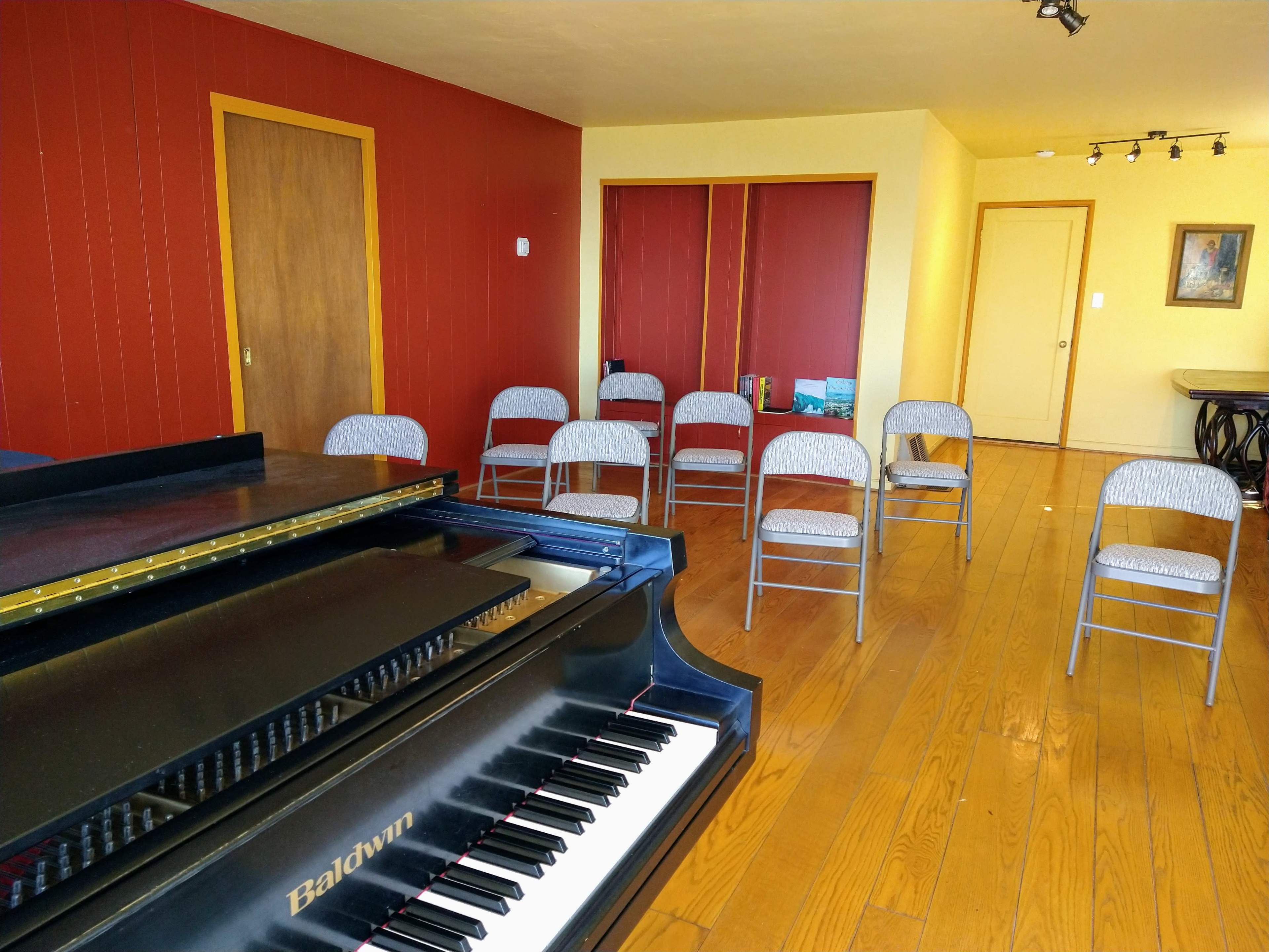 A piano sits in the foreground of a room with wooden floors and several metal chairs arranged in front of it, against a backdrop of red and yellow walls.