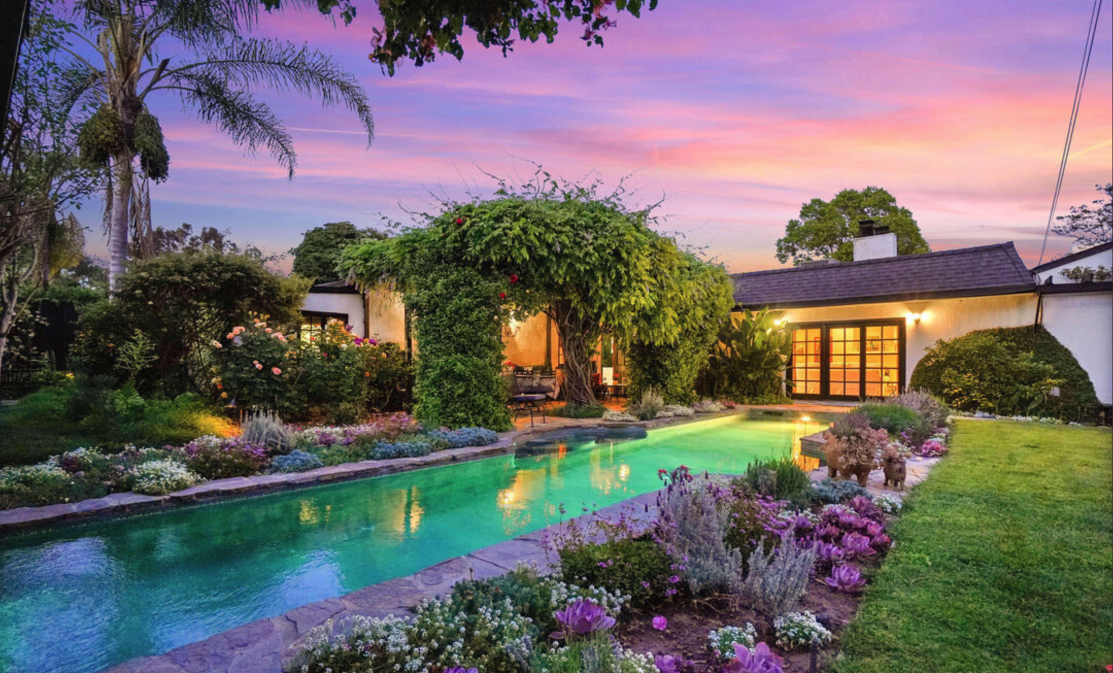 A landscaped backyard features a swimming pool surrounded by colorful flowers and greenery, with a house partially obscured by lush vines under a sunset sky.