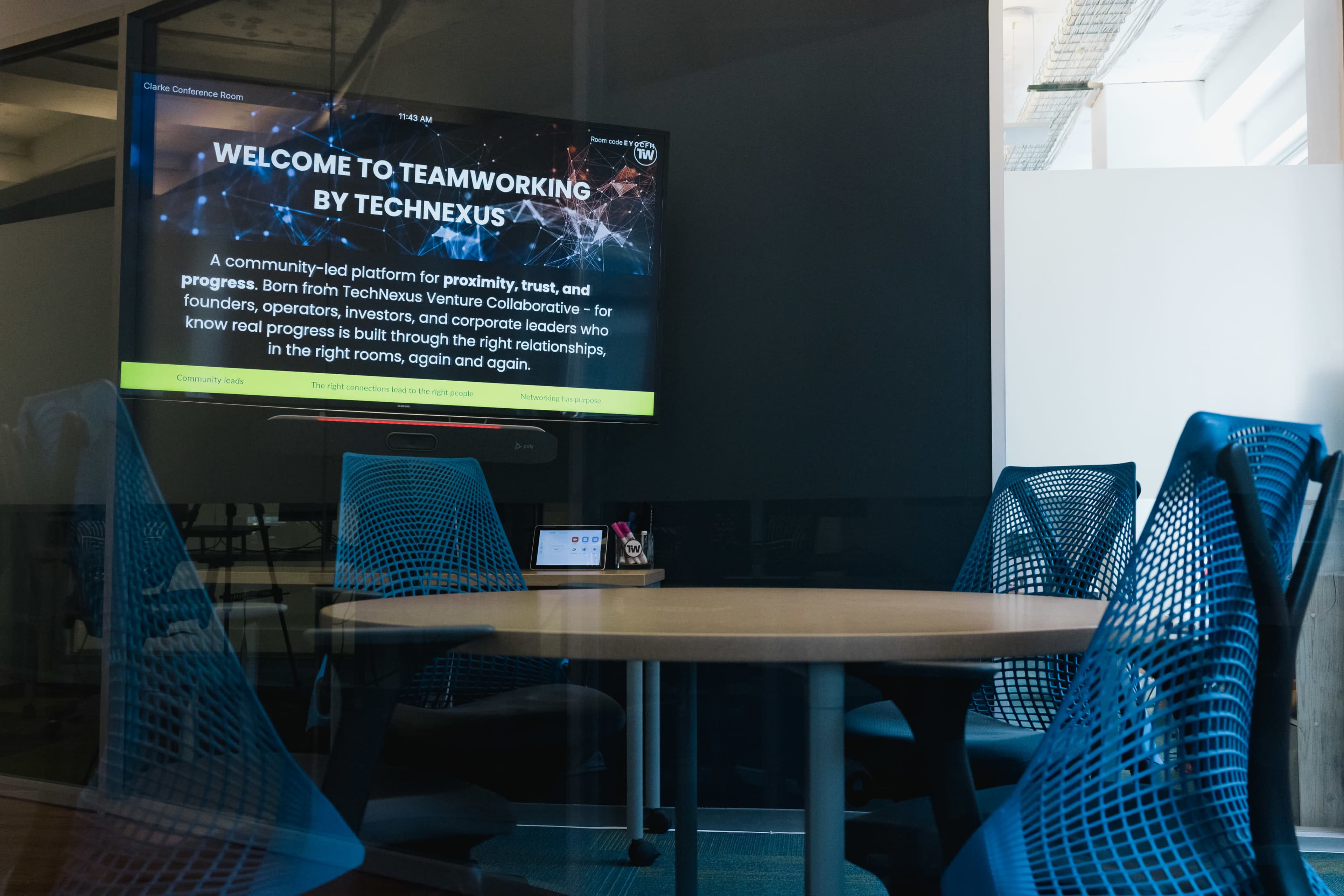The image shows a modern conference room with a circular table and four blue mesh chairs, featuring a screen displaying a welcome message for TechNexus.