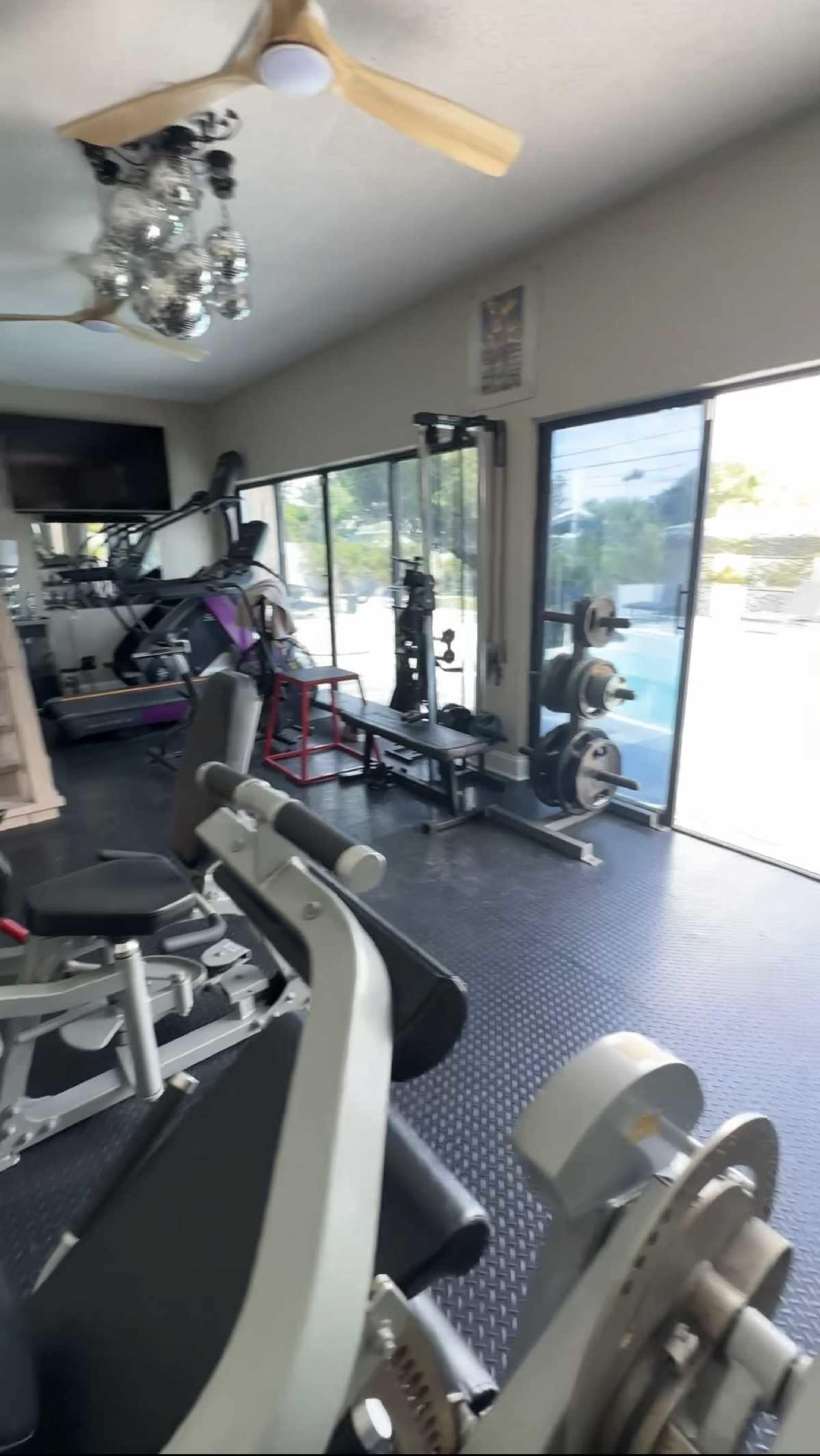 The image shows a fitness room with various exercise equipment, large windows providing natural light, and a pool visible outside.