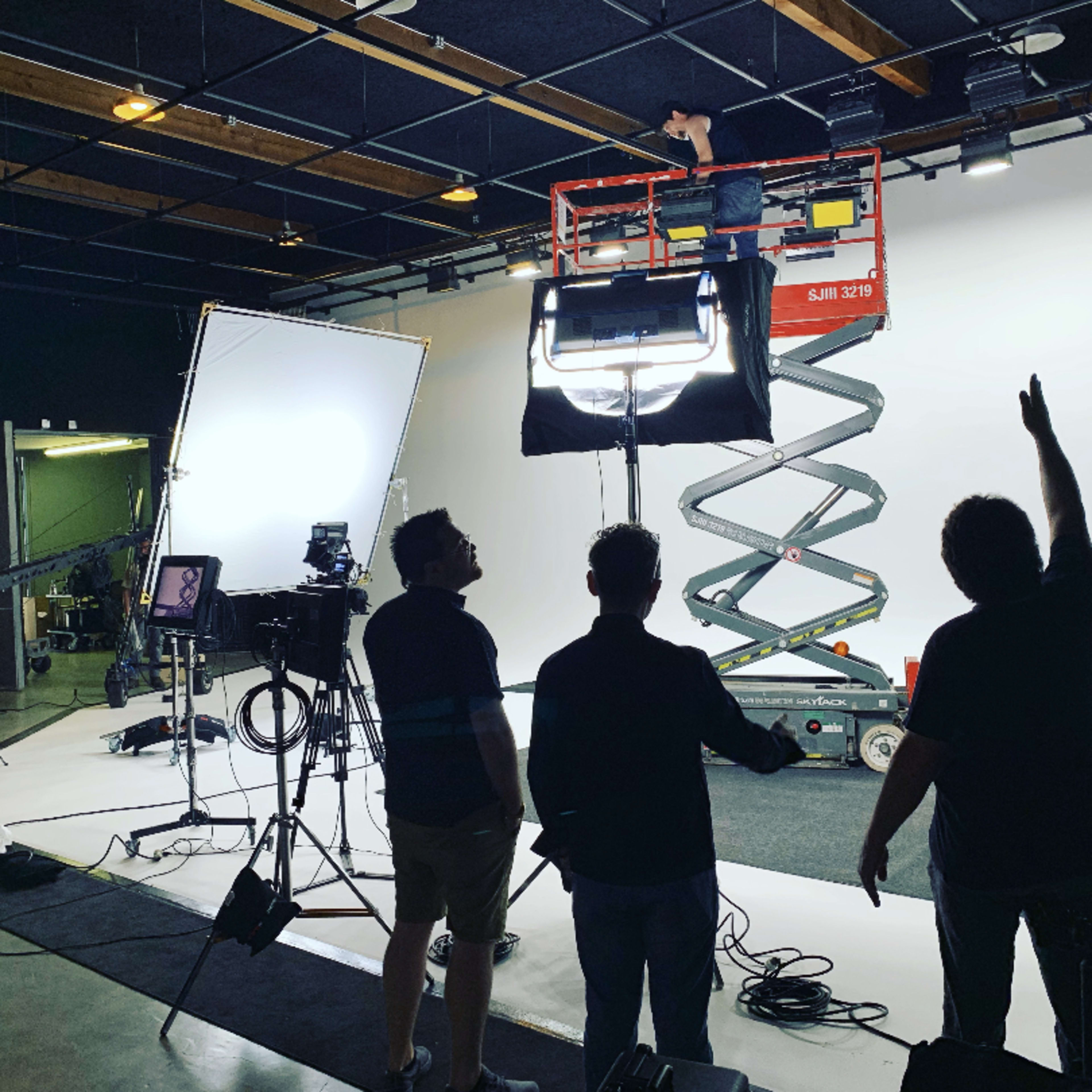 A film crew is setting up equipment in a studio, with one member on a scissor lift adjusting lights while others monitor the scene.