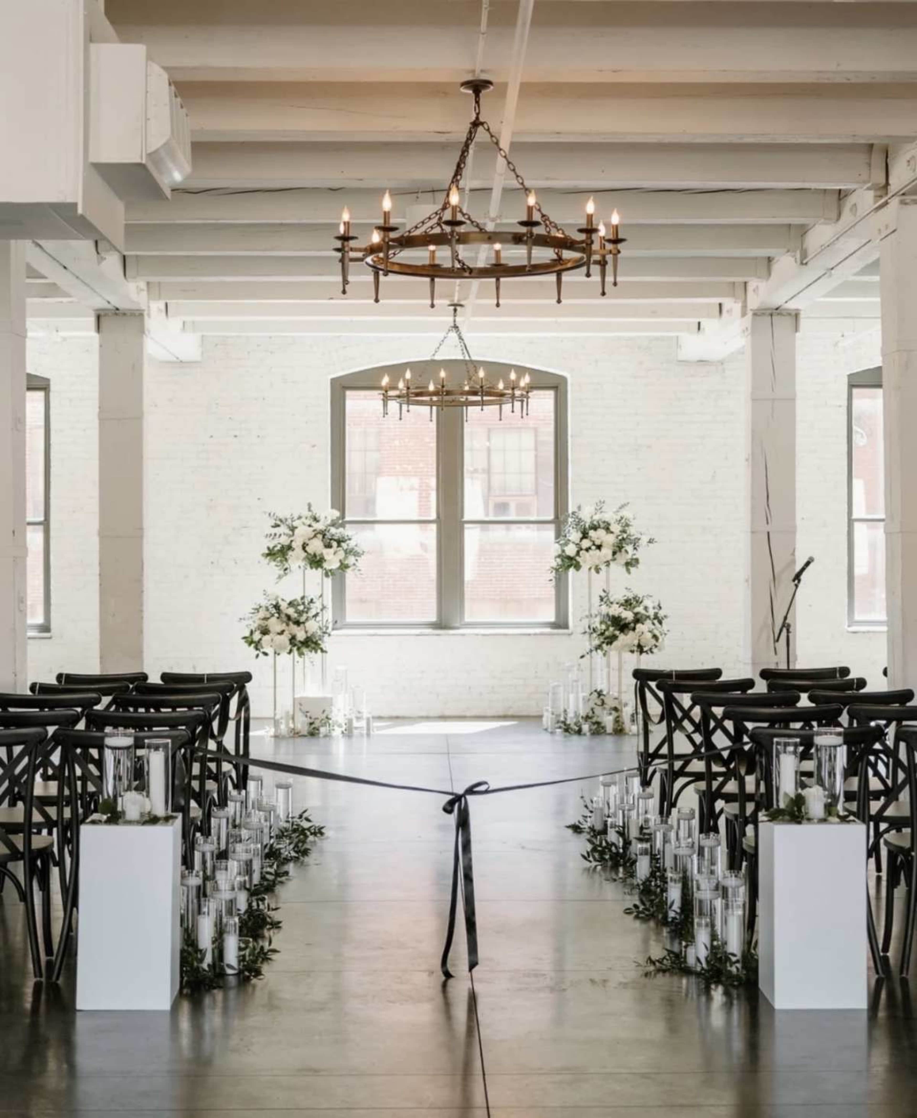 The image shows a wedding ceremony setup in a spacious venue with wooden chairs arranged in rows, flanked by clear candle stands and floral arrangements.