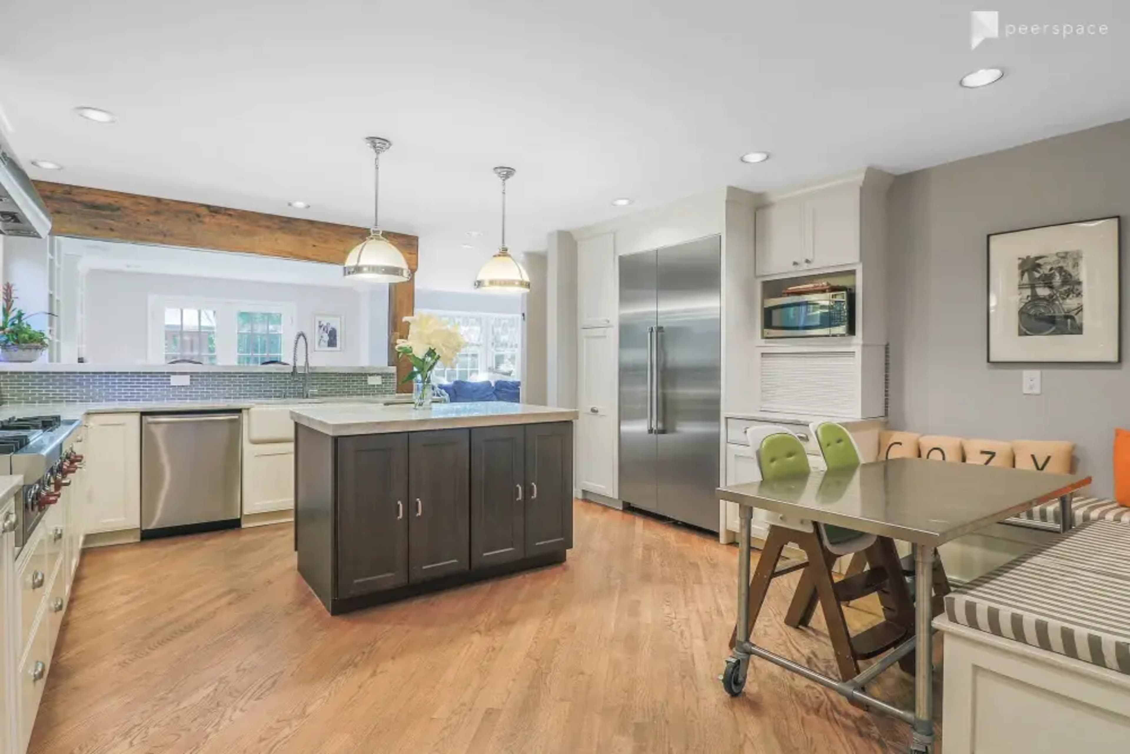 The image shows a modern kitchen featuring a central island with dark cabinetry, stainless steel appliances, and a dining table beside a seating area.