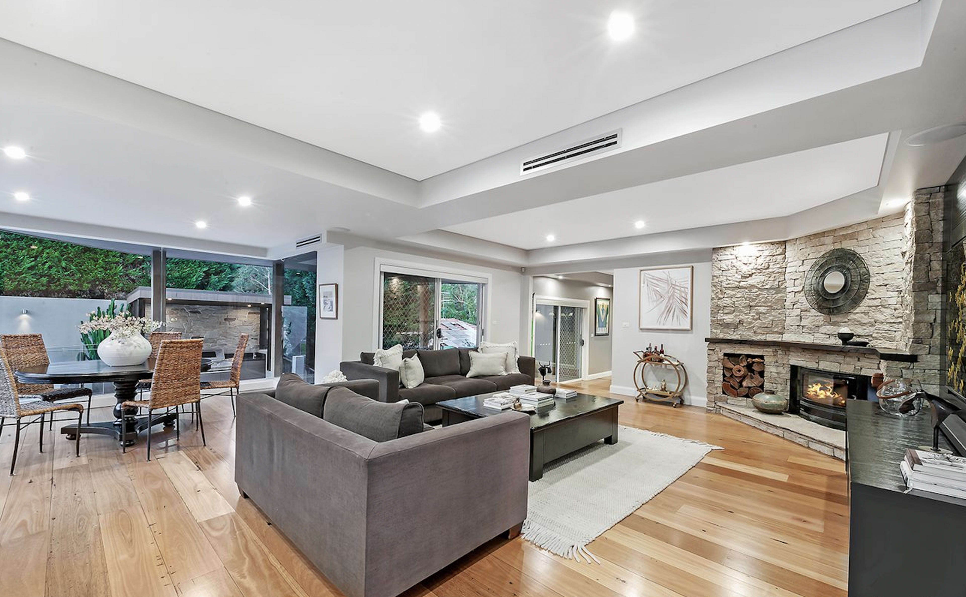 The image shows a spacious living room with a gray sofa, a stone fireplace, and a dining area featuring a glass table, all surrounded by wooden flooring.