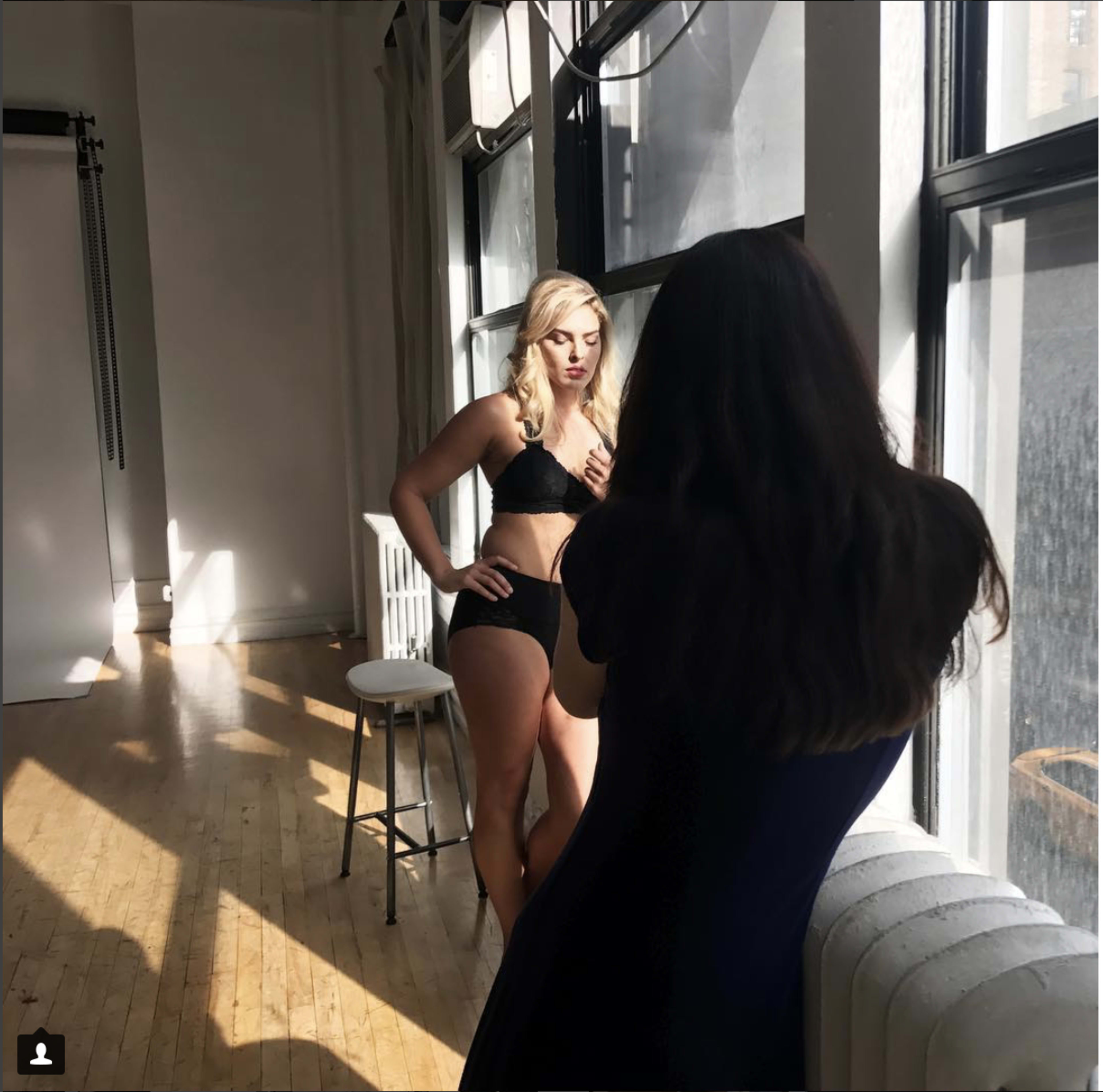 A woman poses in underwear in front of large windows while another woman photographs her.