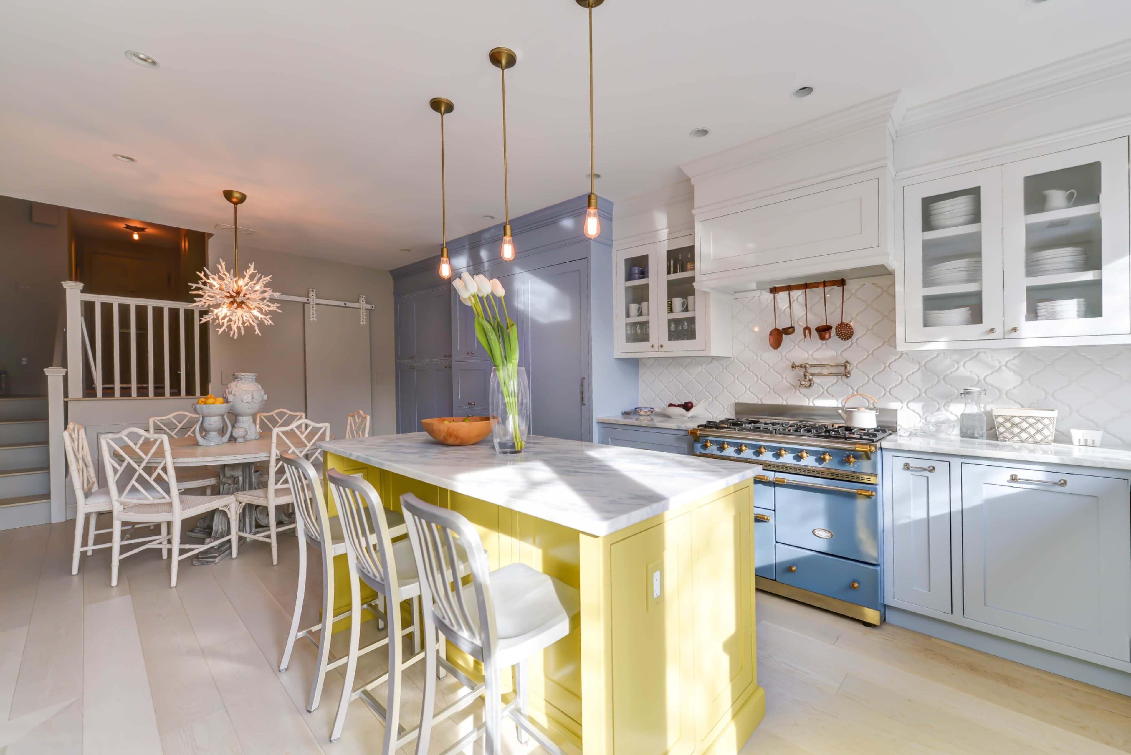 A bright kitchen features a yellow island with white stools, a blue range oven, and a dining area with a round table and decorative light fixture.