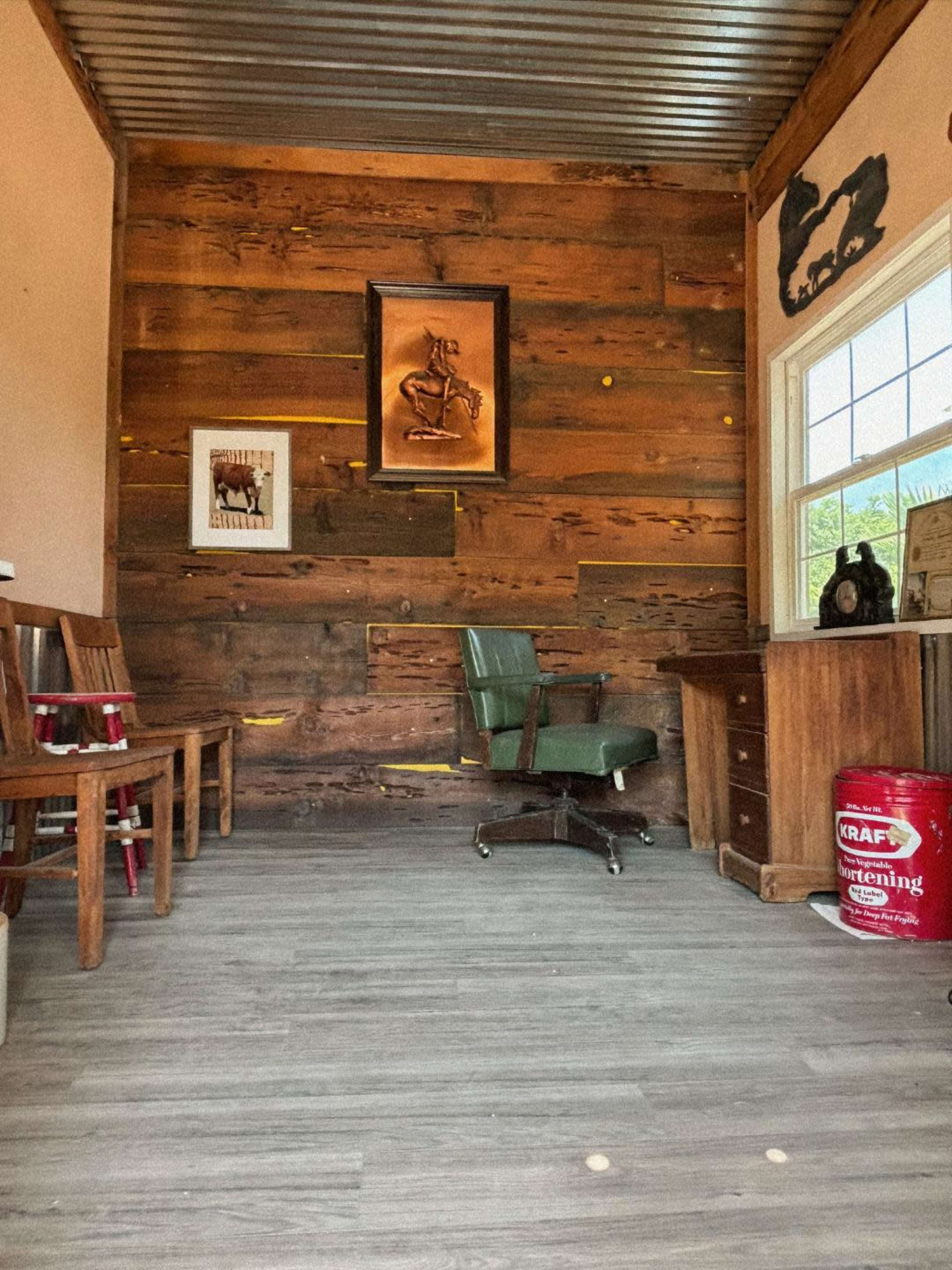 A rustic interior of a room featuring wooden walls, a green office chair, a wooden desk, and various wall decorations, including a framed artwork and a window.