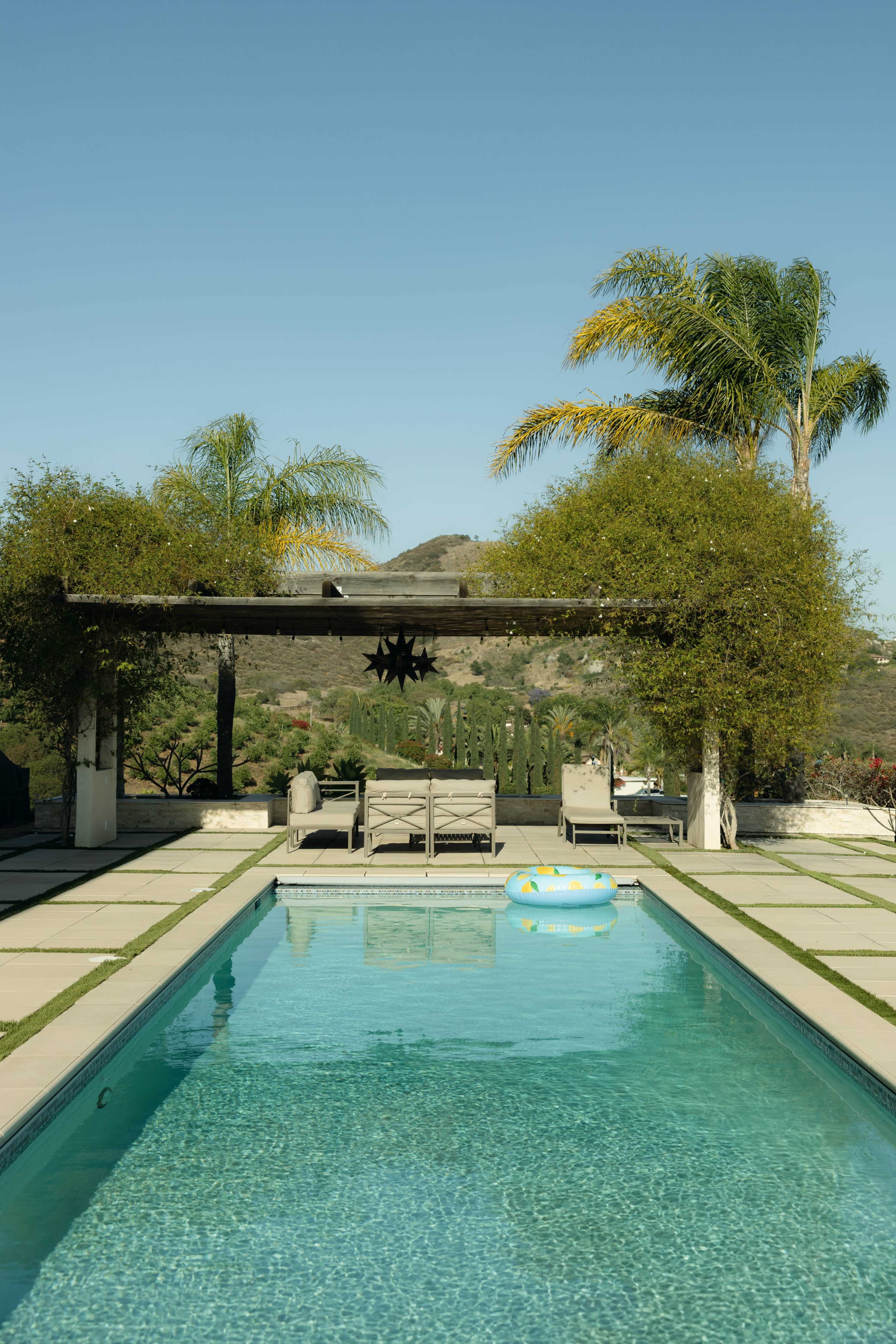 A clear swimming pool is framed by lounge chairs and palm trees, with a backdrop of rolling hills under a blue sky.