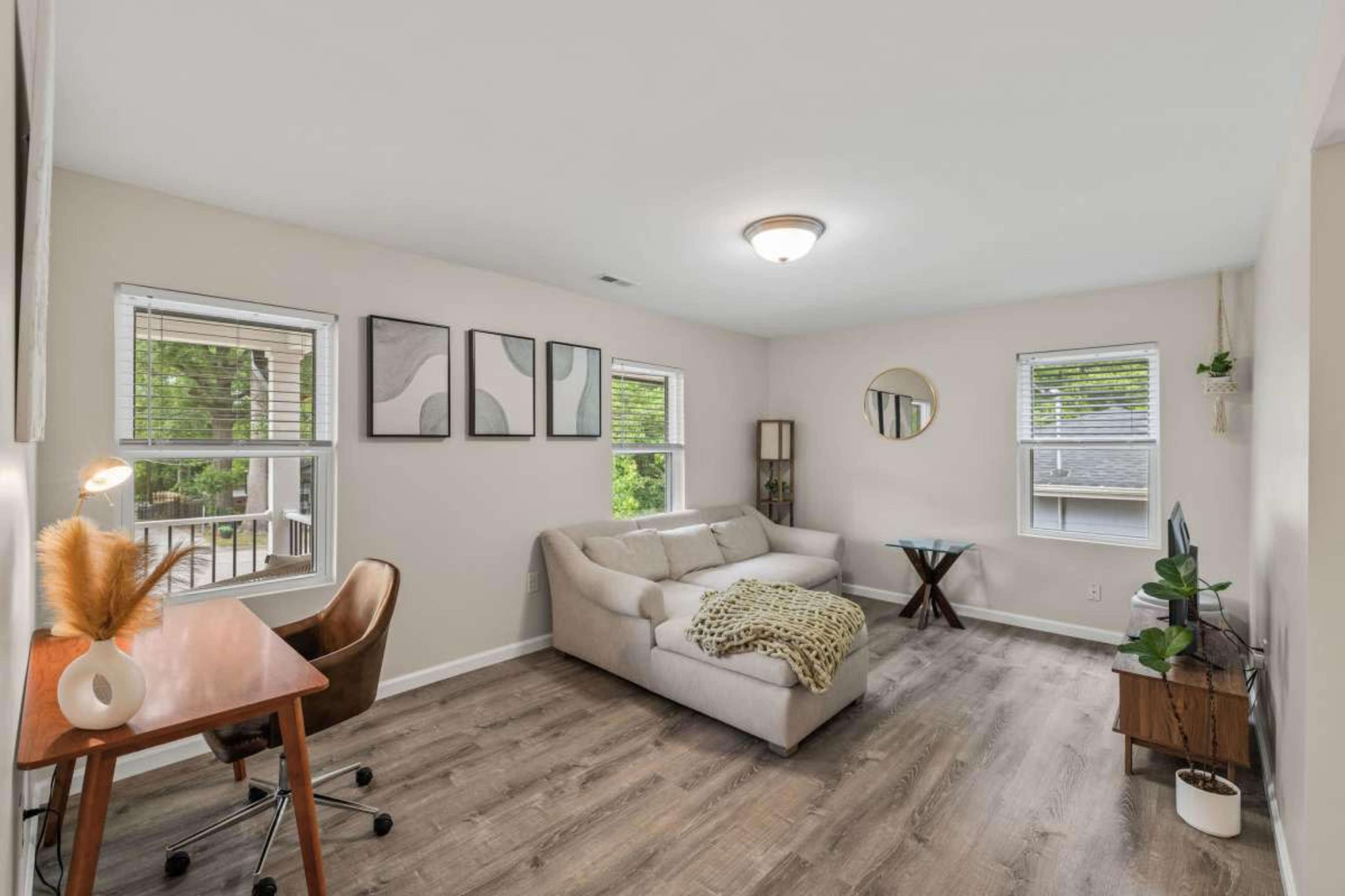 The image depicts a living room with a light-colored sofa, a wooden desk and chair, and several windows allowing natural light to enter the space.