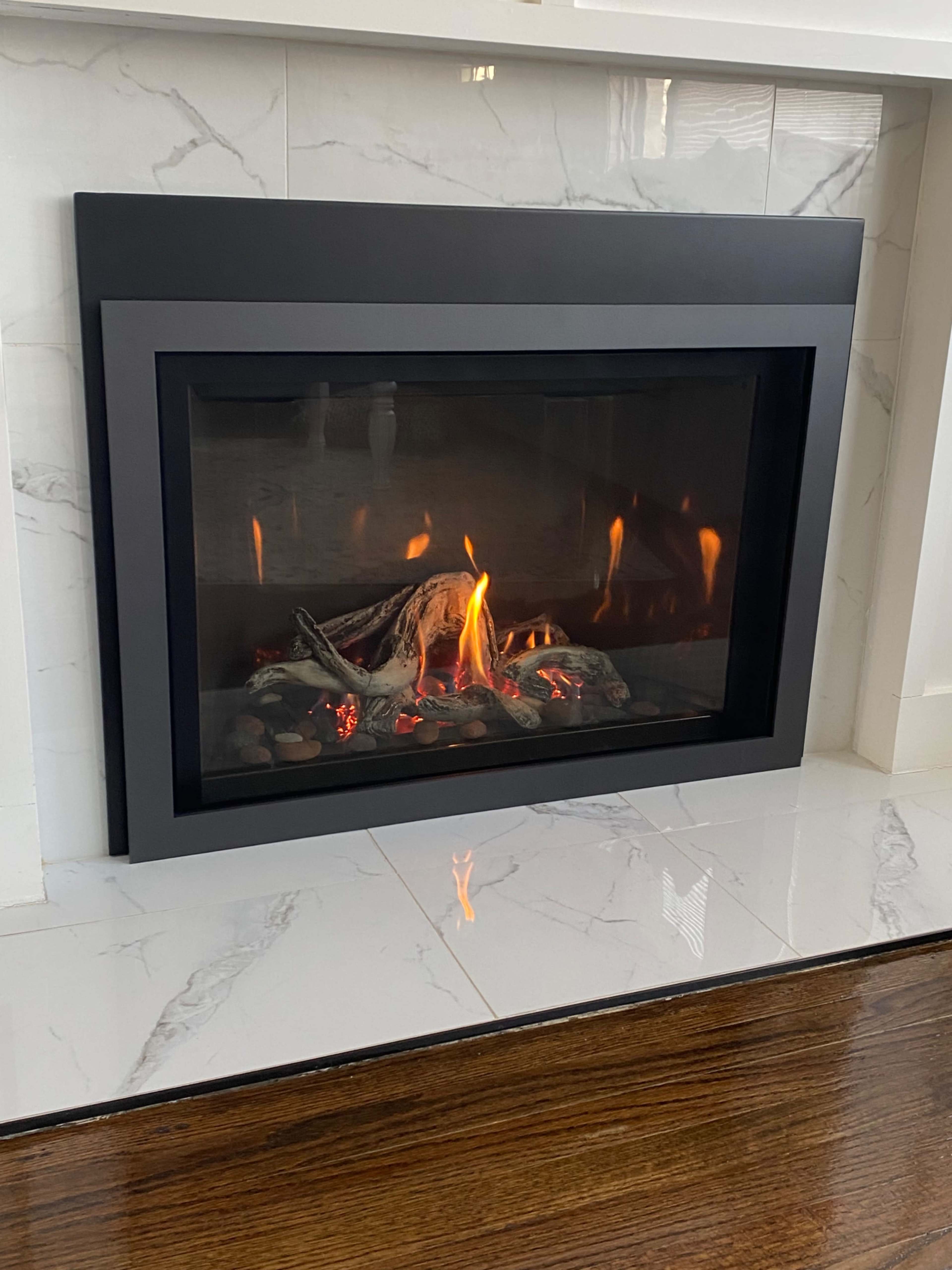 A modern gas fireplace with a glass front is set into a white marble wall, displaying flames and glowing logs on a bed of stones.