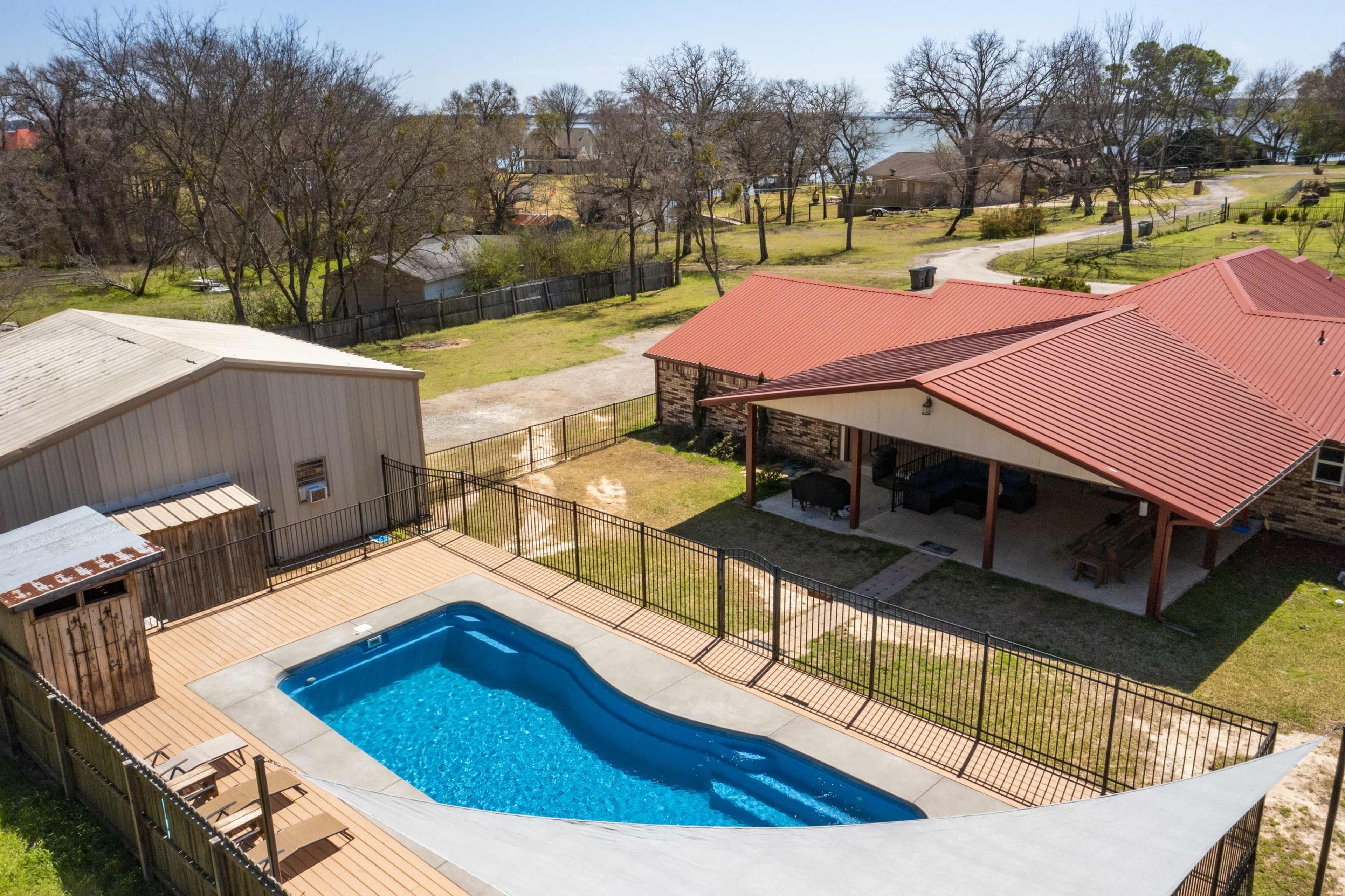 A backyard with a swimming pool surrounded by a fence, a covered patio, and several trees in the background.