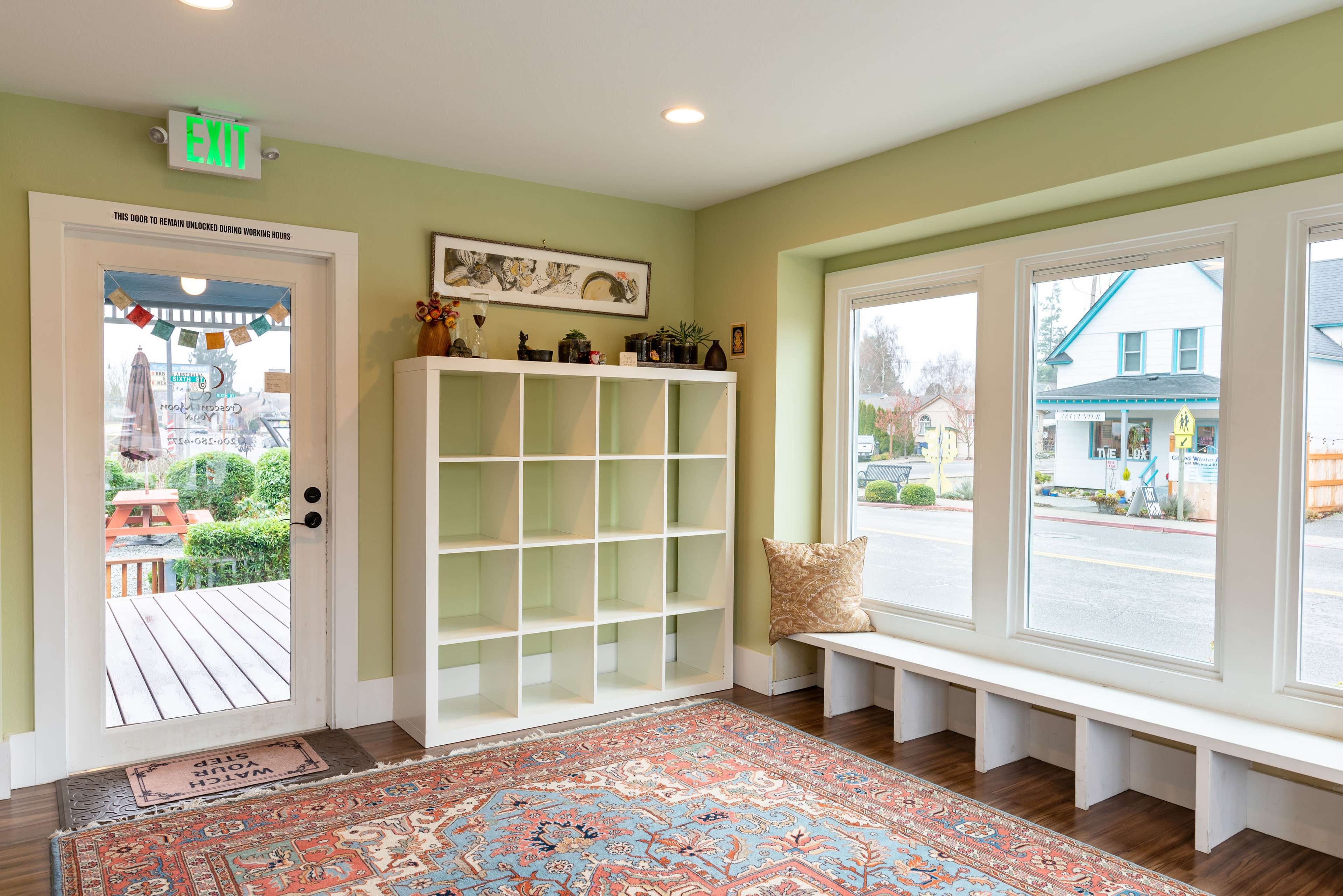The image shows a neatly organized room with green walls, a shelving unit, a bench by the window, and an exit door leading outside.