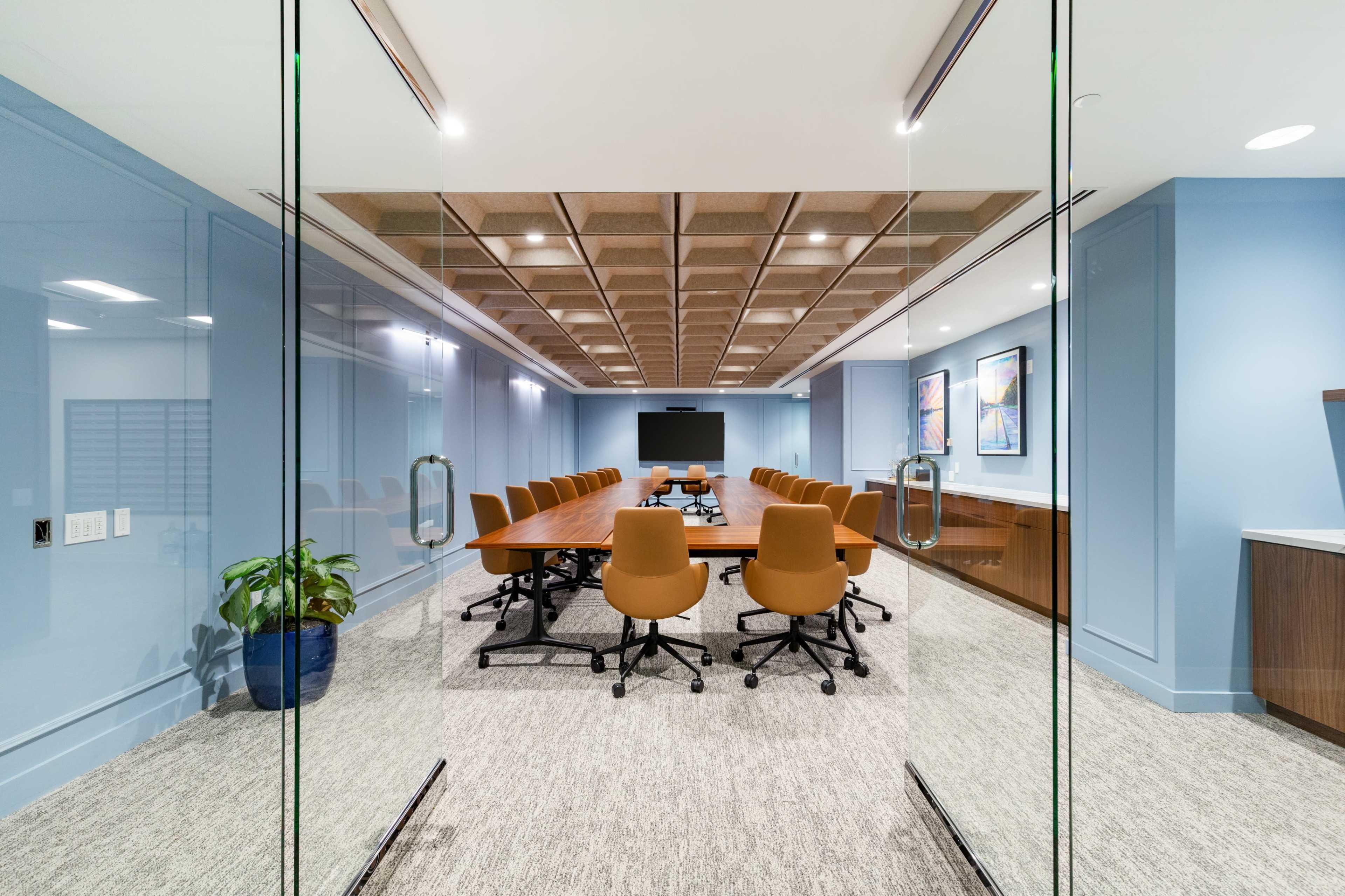 A modern conference room features a long wooden table, ergonomic chairs, and a large screen at the front, framed by blue walls and a coffered ceiling.