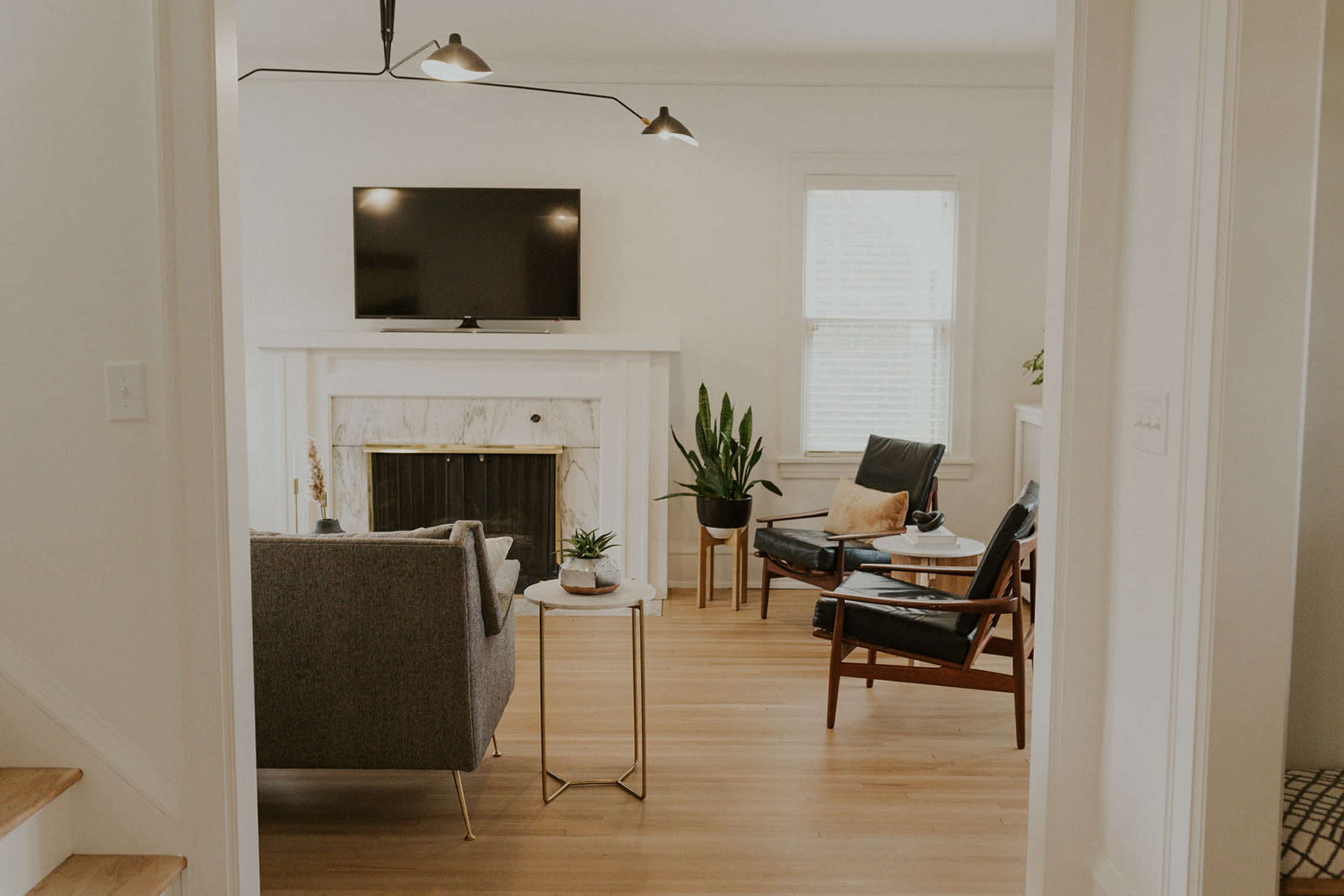 A modern living room features a gray sofa, a wooden chair, a marble fireplace, and a television mounted on the wall.