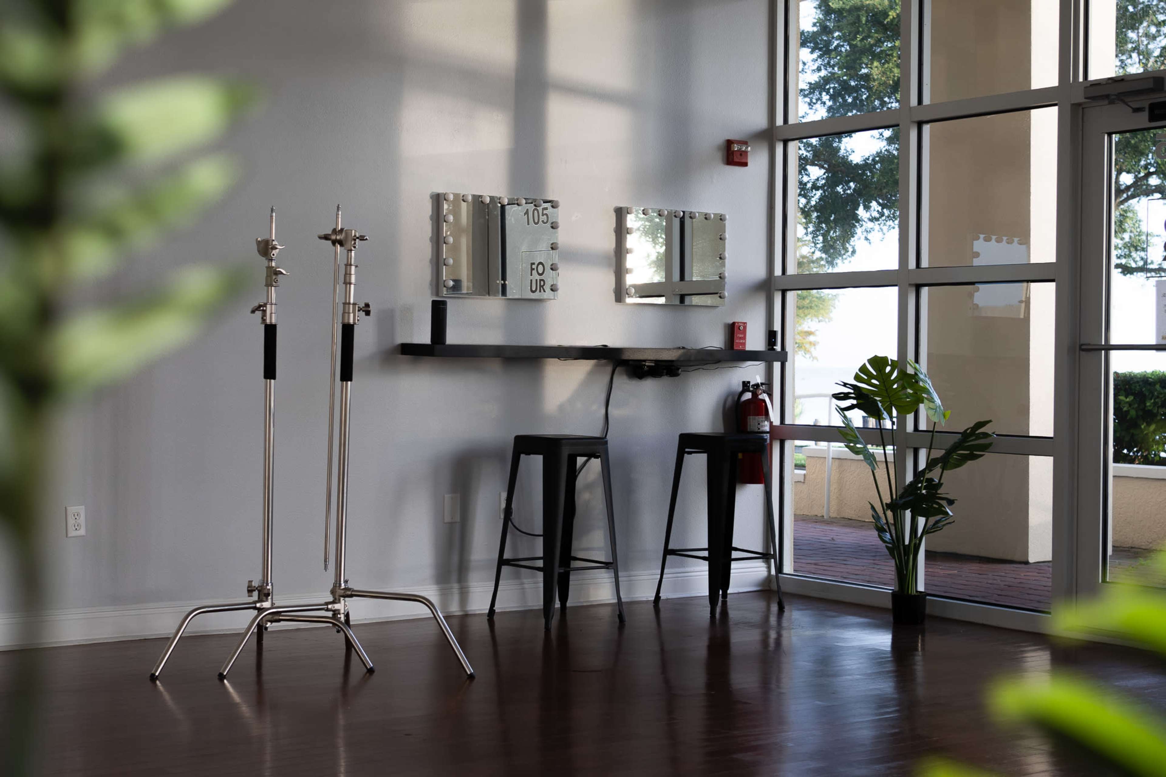 The image shows a modern interior space with a wall of mirrors, two stools, and a clothing rack, illuminated by natural light from large windows.